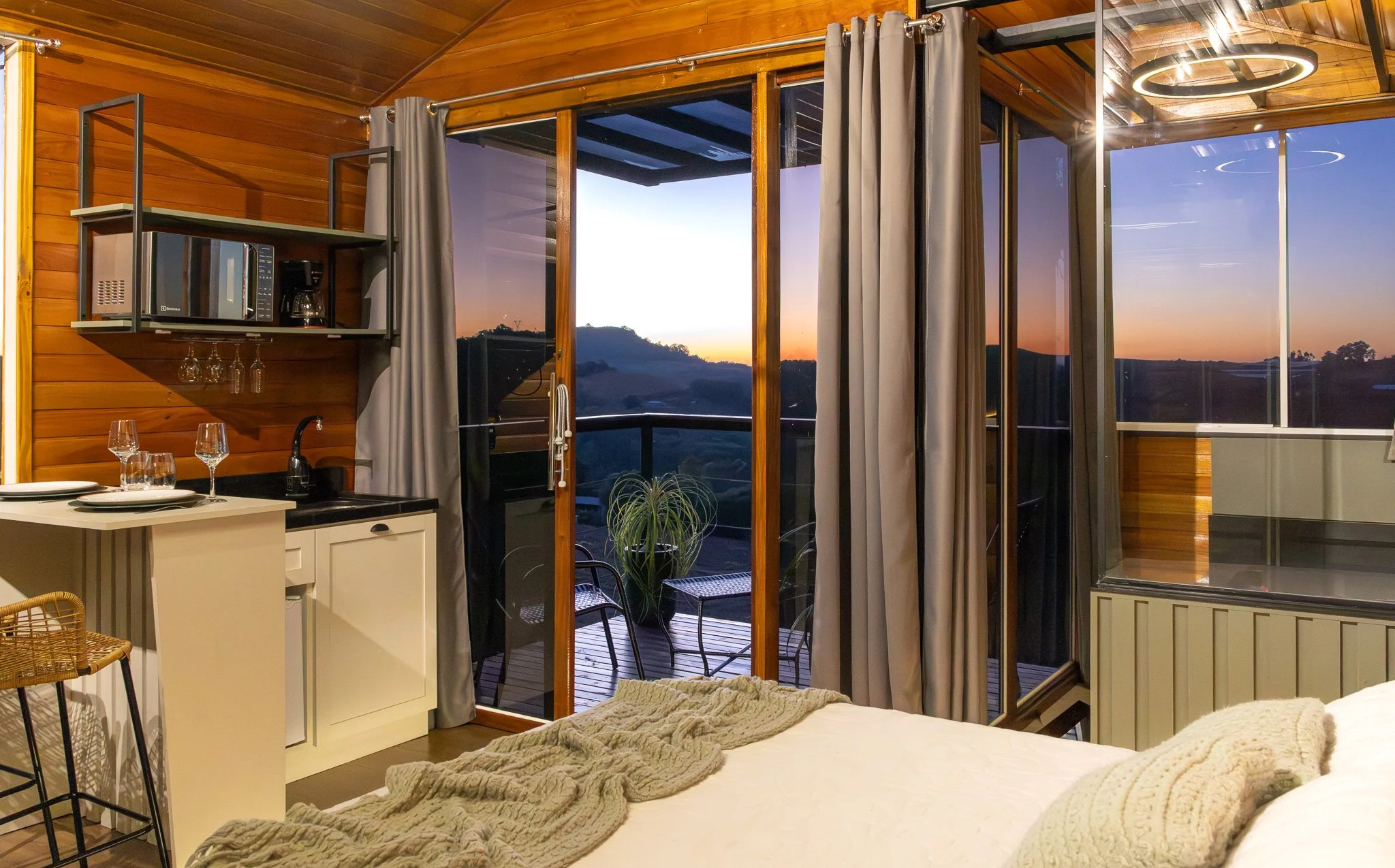 Vista de um quarto de uma casa de madeira com portas de correr de vidro que dão para uma varanda com vista para o pôr do sol ou nascer do sol com colinas ao fundo.