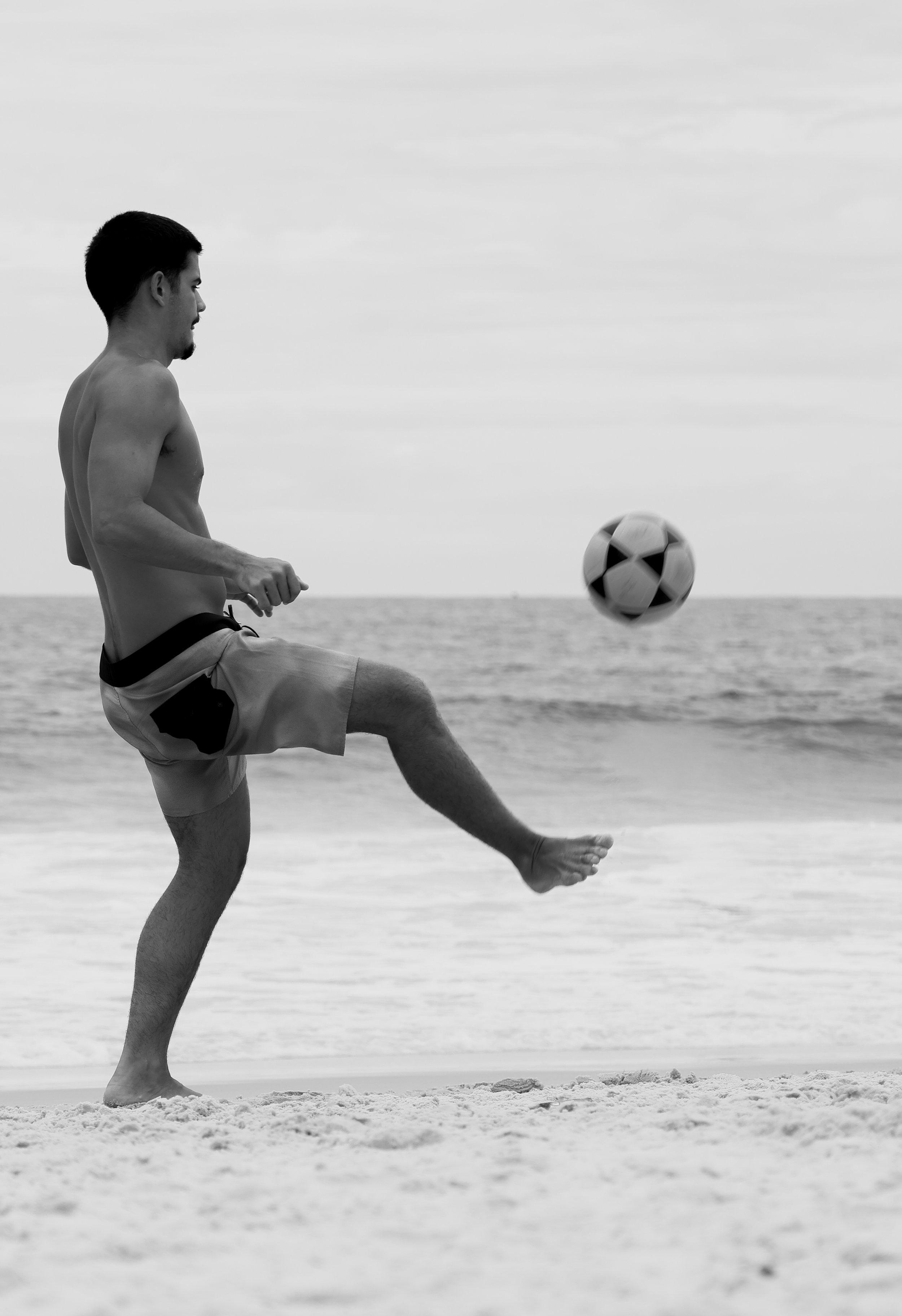Homem jogando bola de futebol na praia, vestindo shorts, em um ambiente ao ar livre