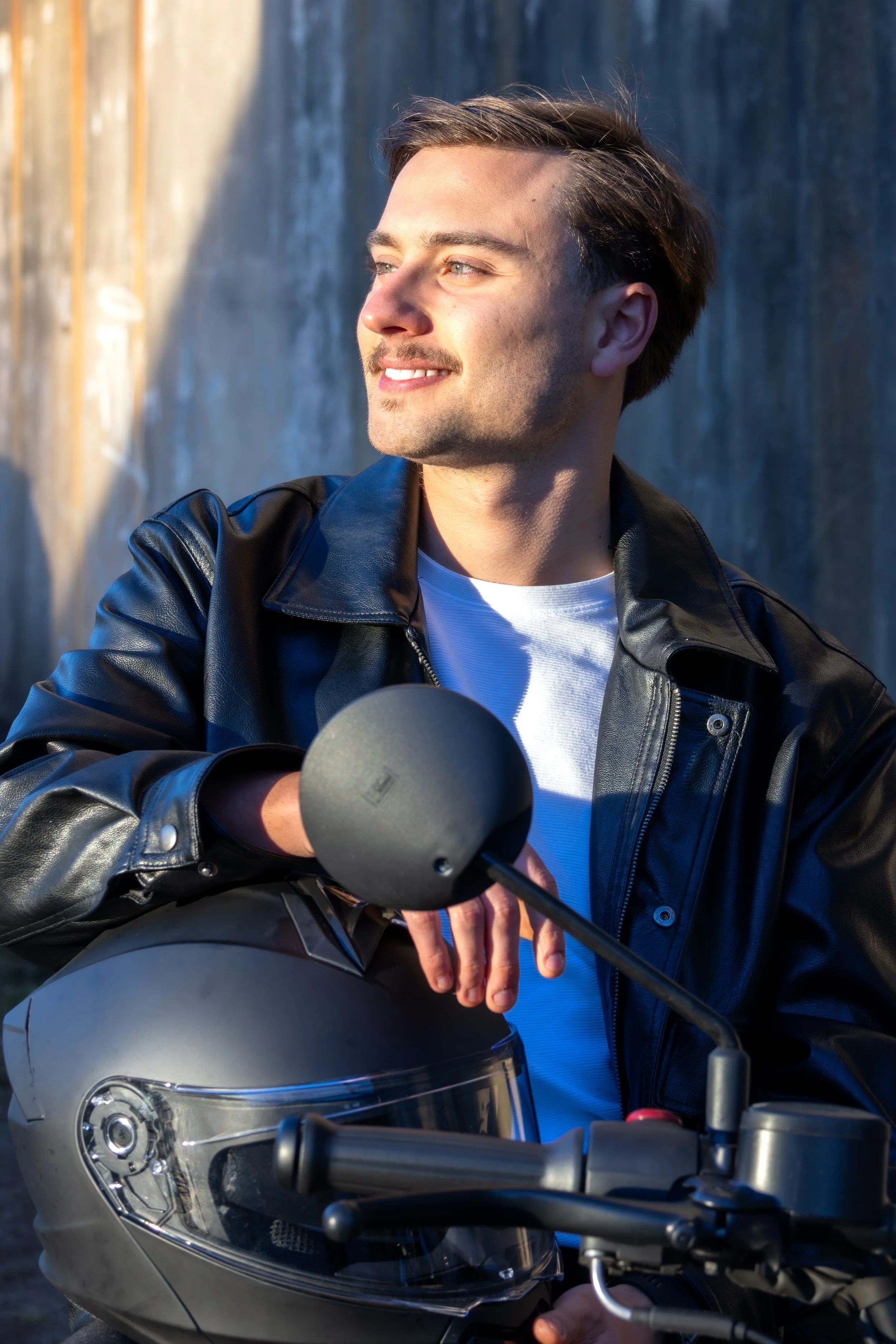 Homem sorrindo sentado em uma moto com capacete na mão, usando jaqueta de couro e camiseta branca, em ambiente ao ar livre com parede ao fundo.