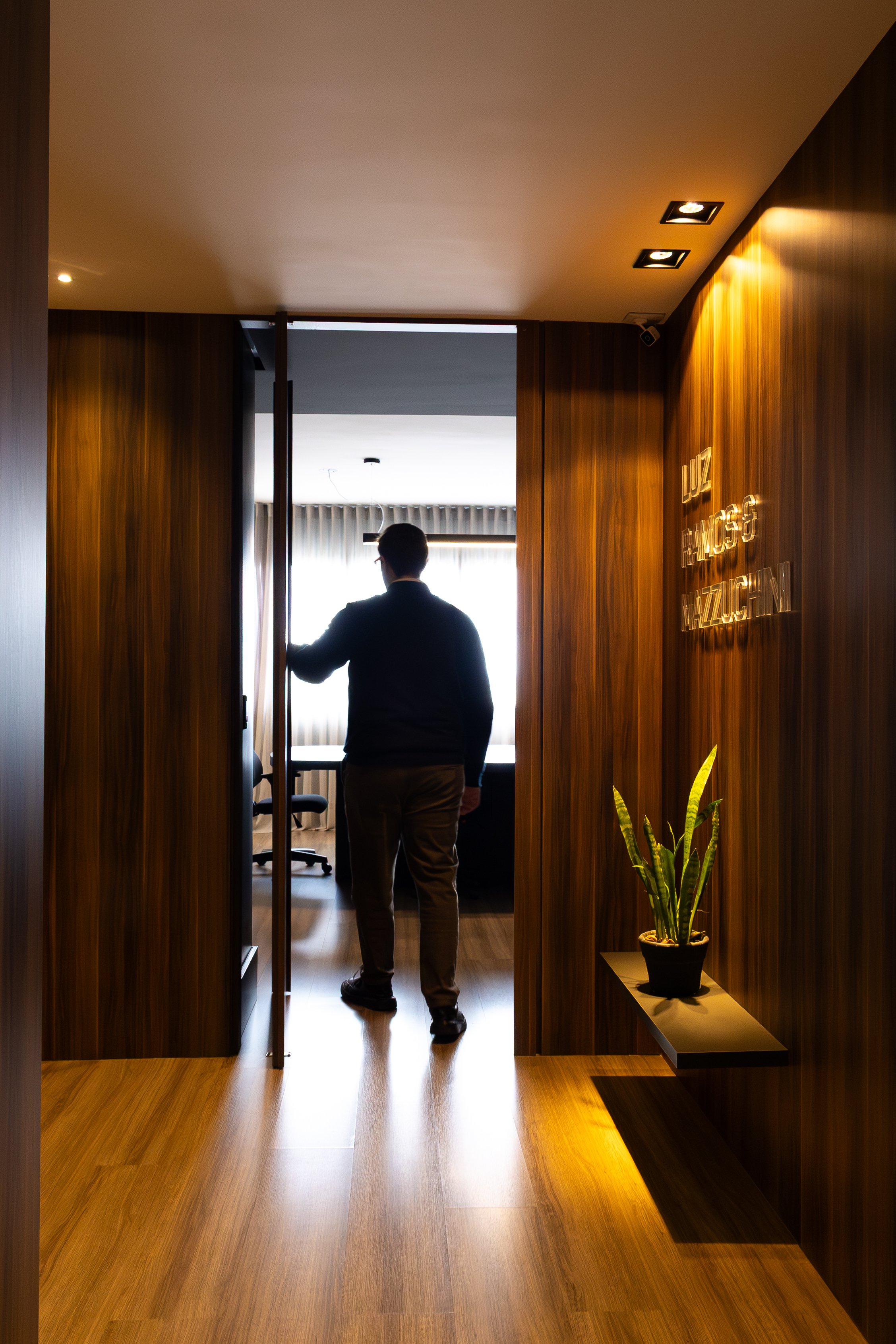 Pessoa de costas segurando uma porta de vidro no interior de um escritório com paredes de madeira, decorado com plantas e iluminação suave.