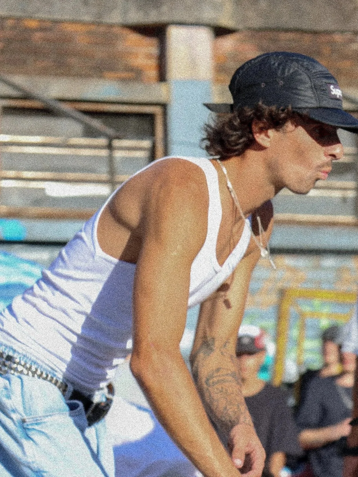 Pessoa jovem com cabelo cacheado, usando boné preto, camiseta branca, e com tatuagens nos braços, posando ao ar livre em um dia ensolarado.