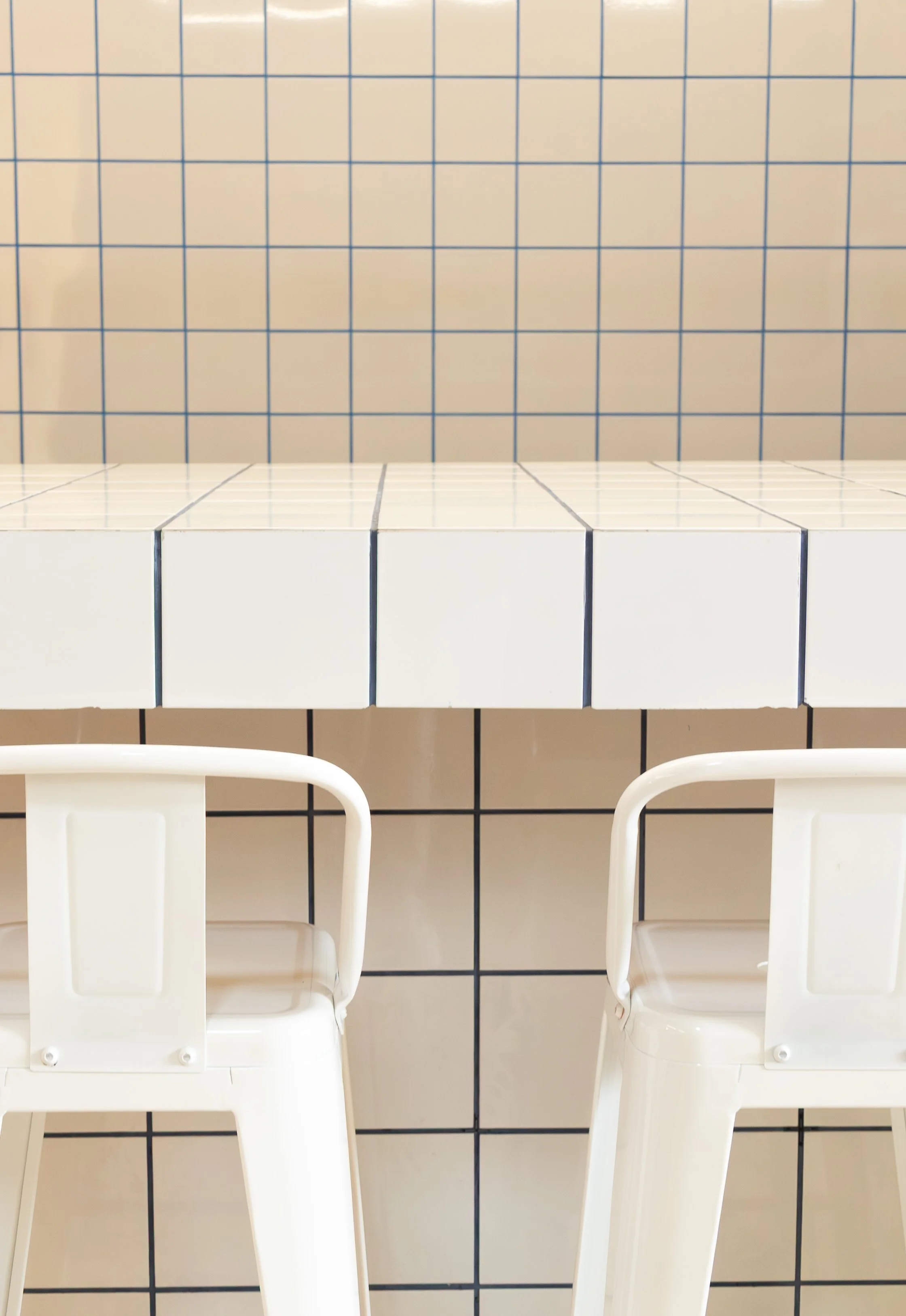 Duas cadeiras brancas de plástico em frente a uma bancada de azulejos branca com linhas pretas, parede com azulejos bege com linhas azuis, no fundo.