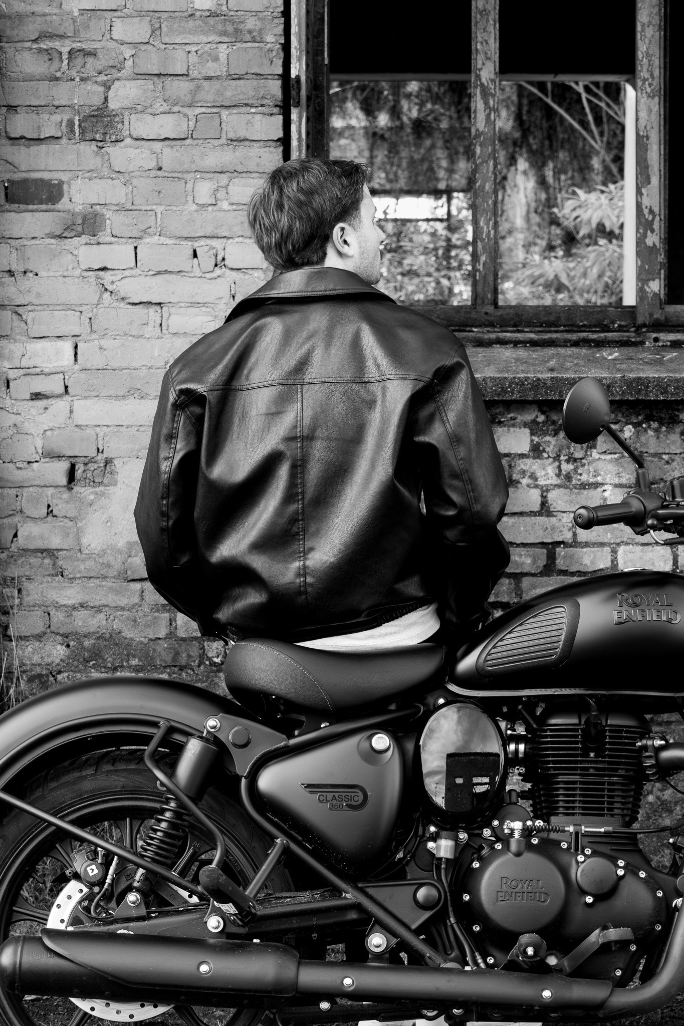 Homem de costas usando jaqueta de couro, sentado em uma scooter preta com logo 'Royal Enfield', diante de uma parede de tijolos e uma janela antiga. sensação de urbanidade e estilo clássico.