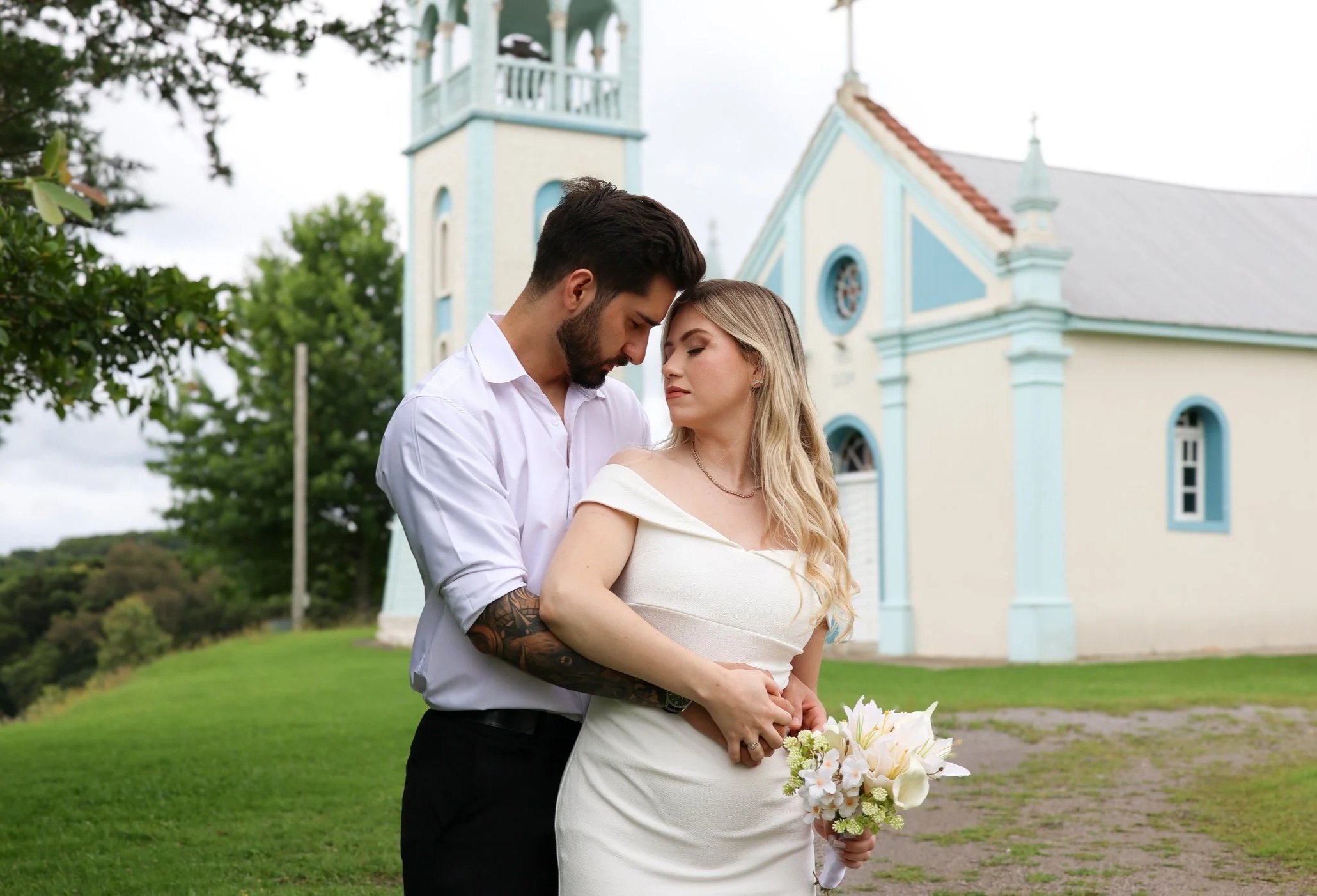 Casal de noivos em abraço diante de uma igreja branca com detalhes azuis, em um dia nublado, segurando buquê de flores brancas.