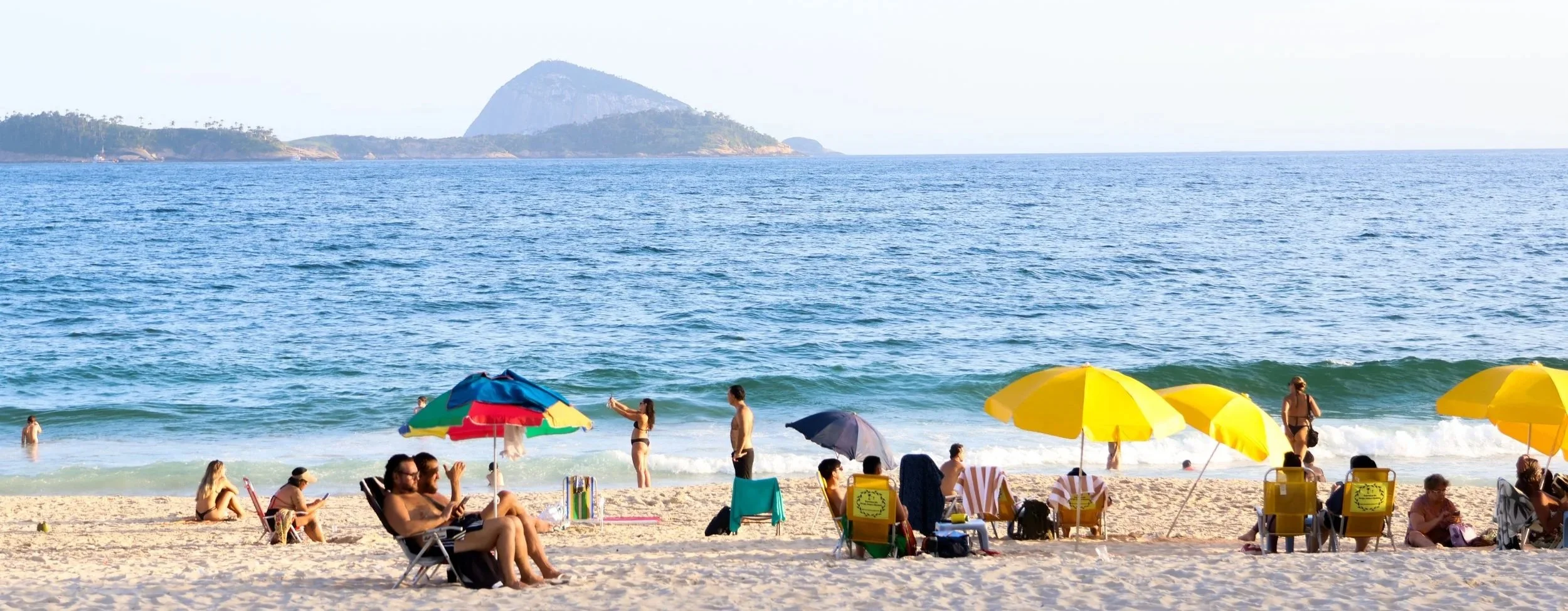 Praia com banhistas, guarda-sóis amarelos e uma barraca de praia. Ilha ao fundo no oceano.