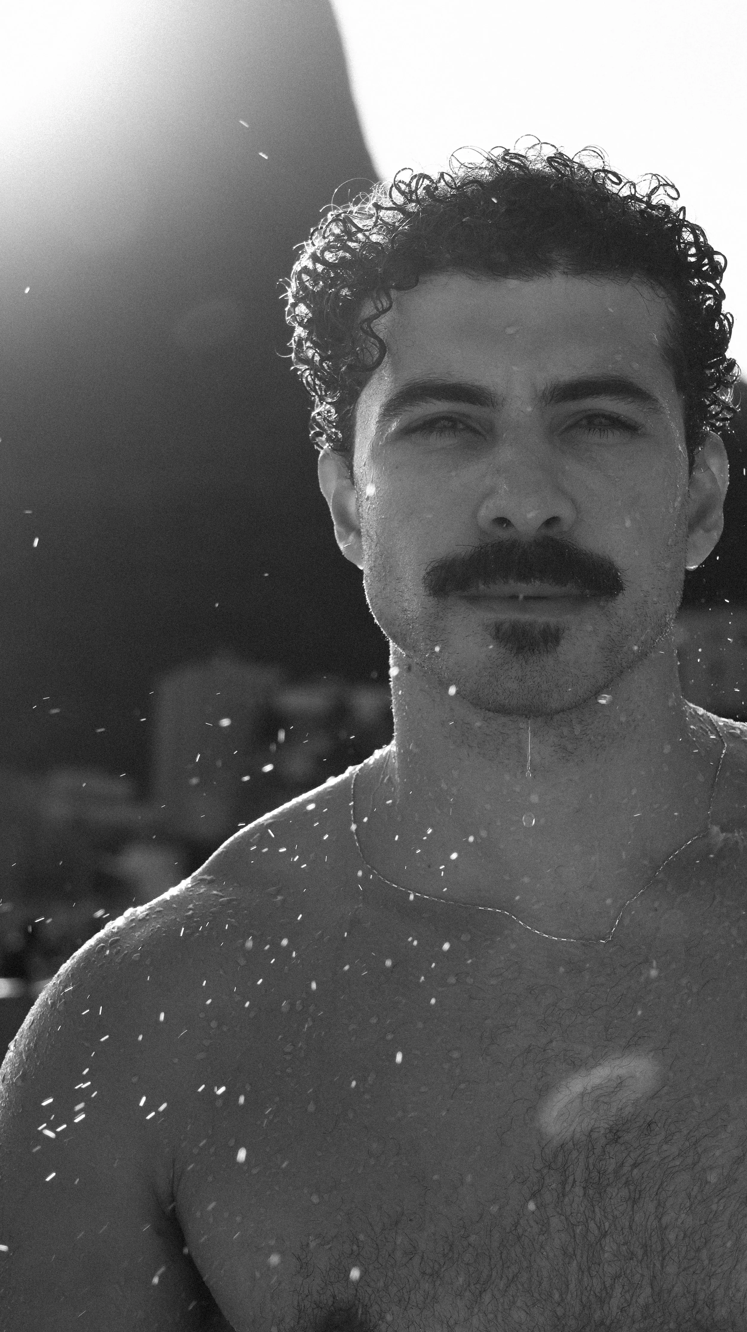 Homem com barba e cabelo cacheado, com gotas d'água no corpo, olhando para a câmera, ao ar livre, em preto e branco.