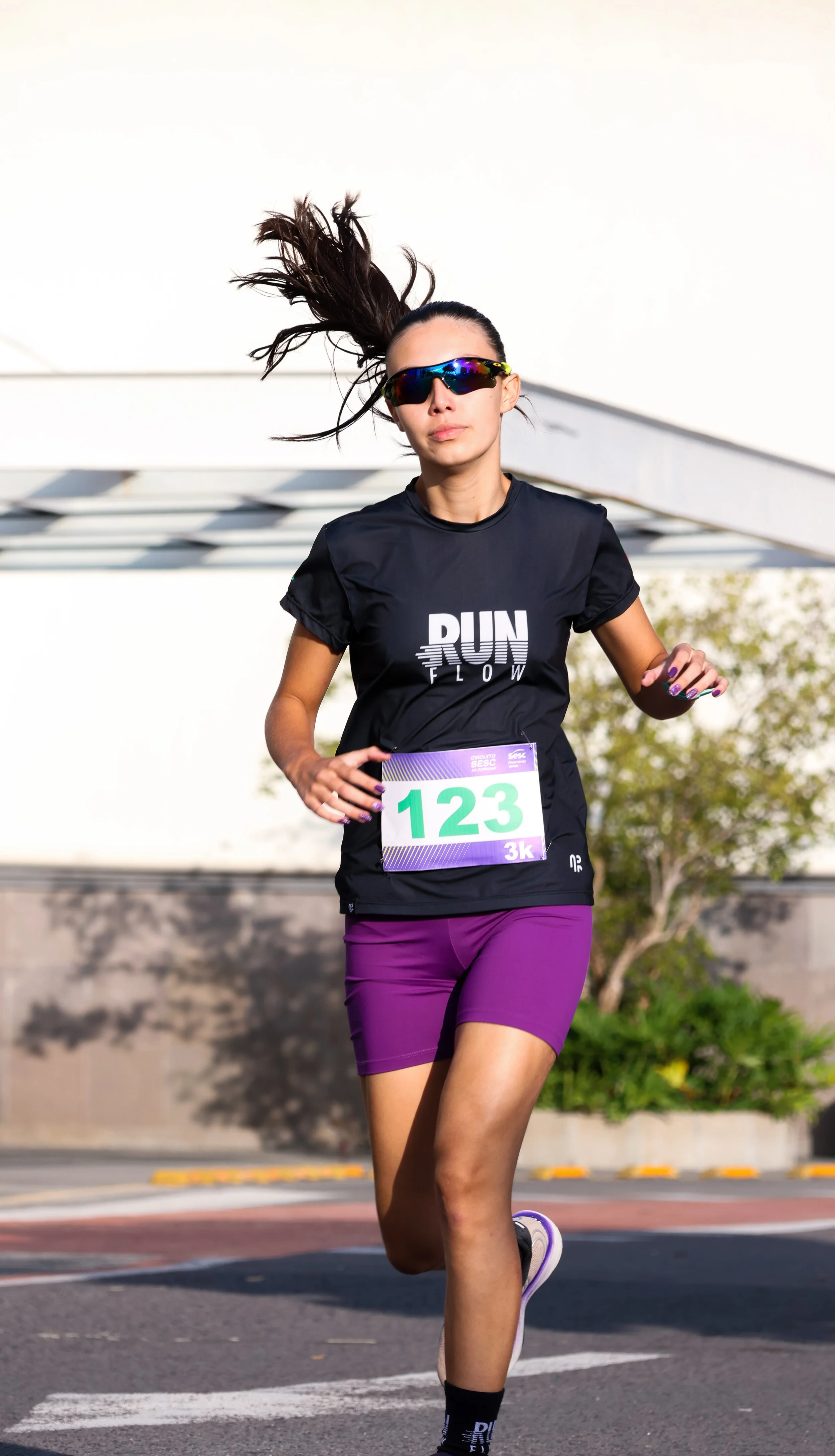 Mulher corredora com camiseta preta que diz "RUN FLOWER", usando óculos escuros esportivos, shorts roxos, com número de corrida 123 na frente, em uma corrida ao ar livre