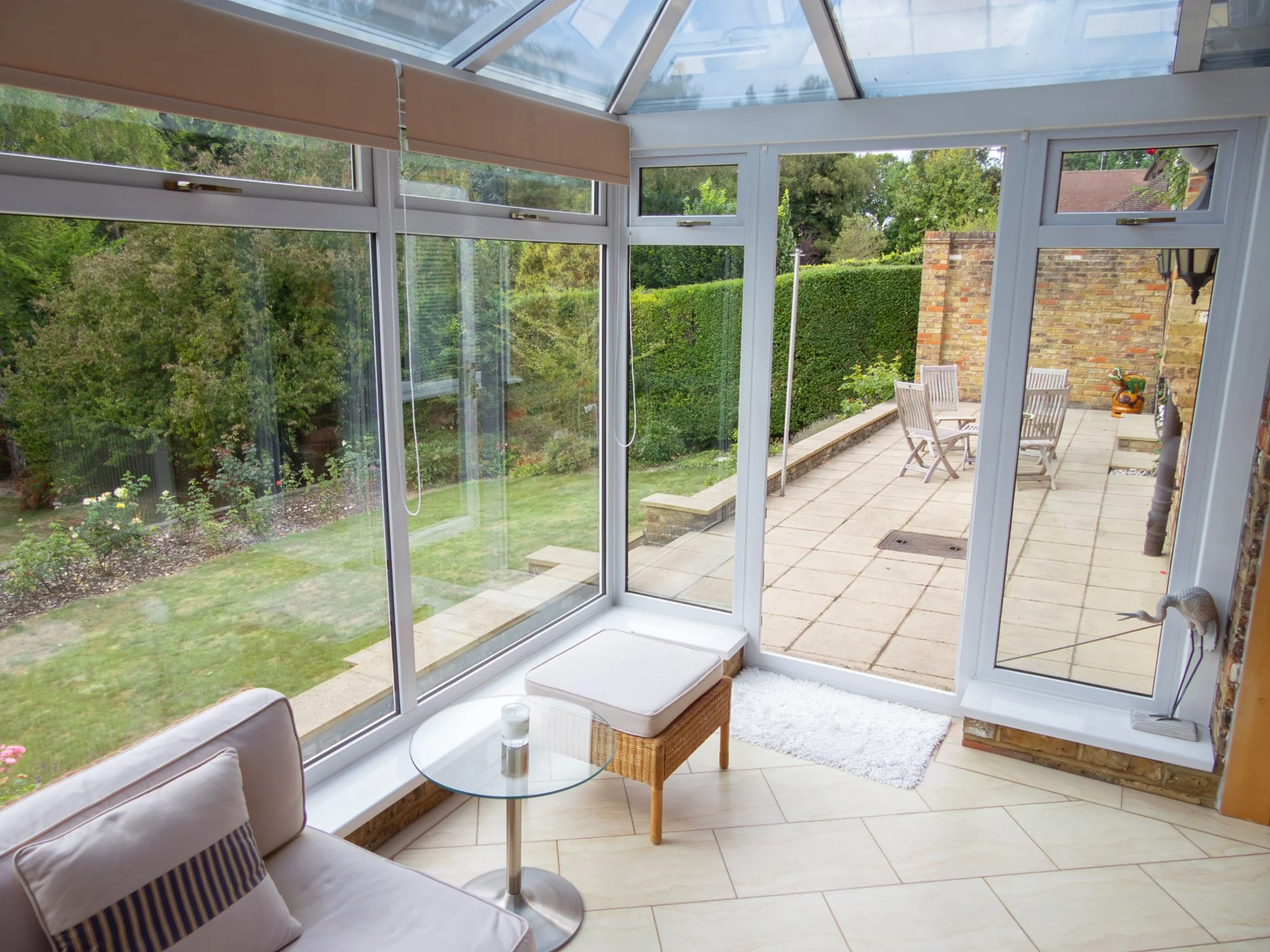 A sunroom with large glass windows looking out to a backyard with green trees and a patio with outdoor furniture.