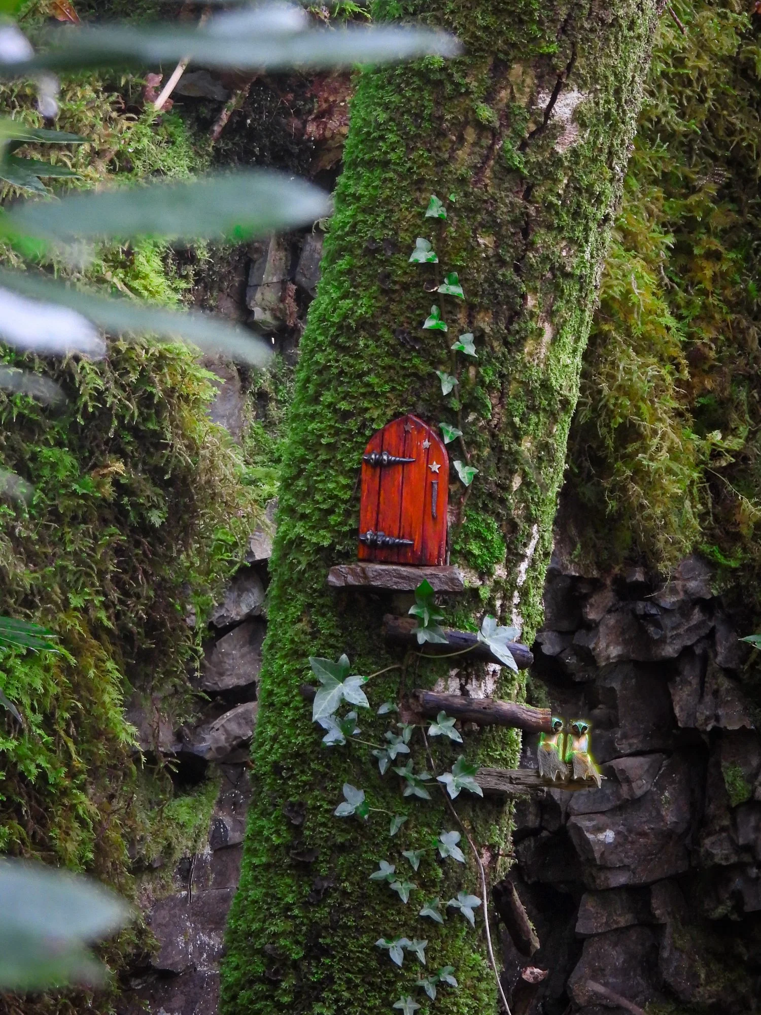 Moss covered tree, wooden steps attached to bark leading to a fantasy red door, two fairies sitting on the bottom step, ivy growing up the tree.