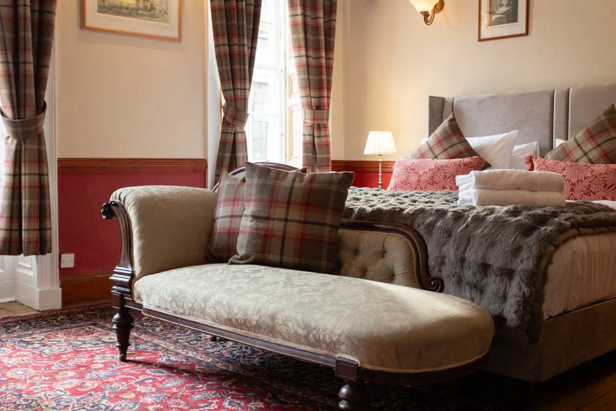 A cozy bedroom with a large bed decorated in plaid pillows and a plush fur throw. A vintage-style chaise lounge sits at the foot of the bed, with matching plaid pillows. The room has patterned curtains, a small lamp on a bedside table, and framed artwork on the walls. The floor features a detailed patterned rug.