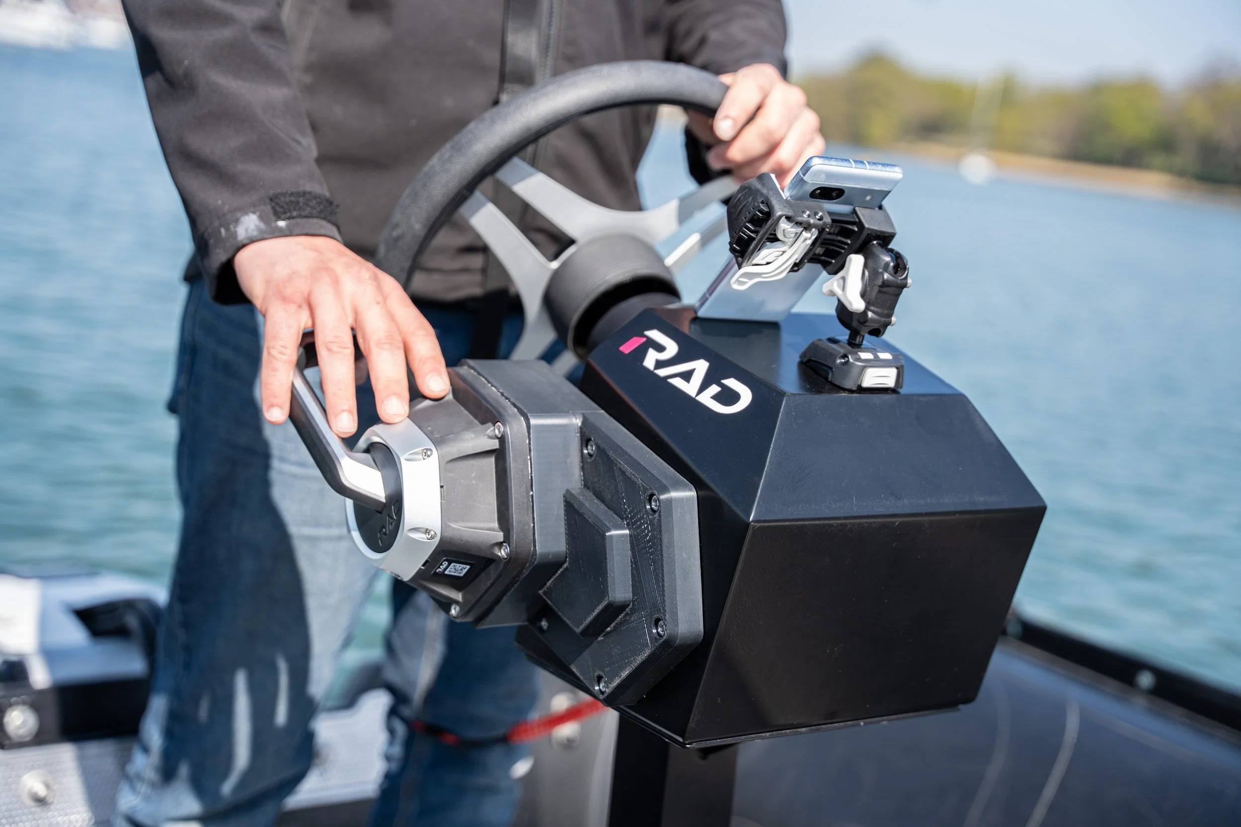 Close-up of a person operating a RIB or small watercraft with a control panel and smartphone mounted on the steering wheel by a lake.