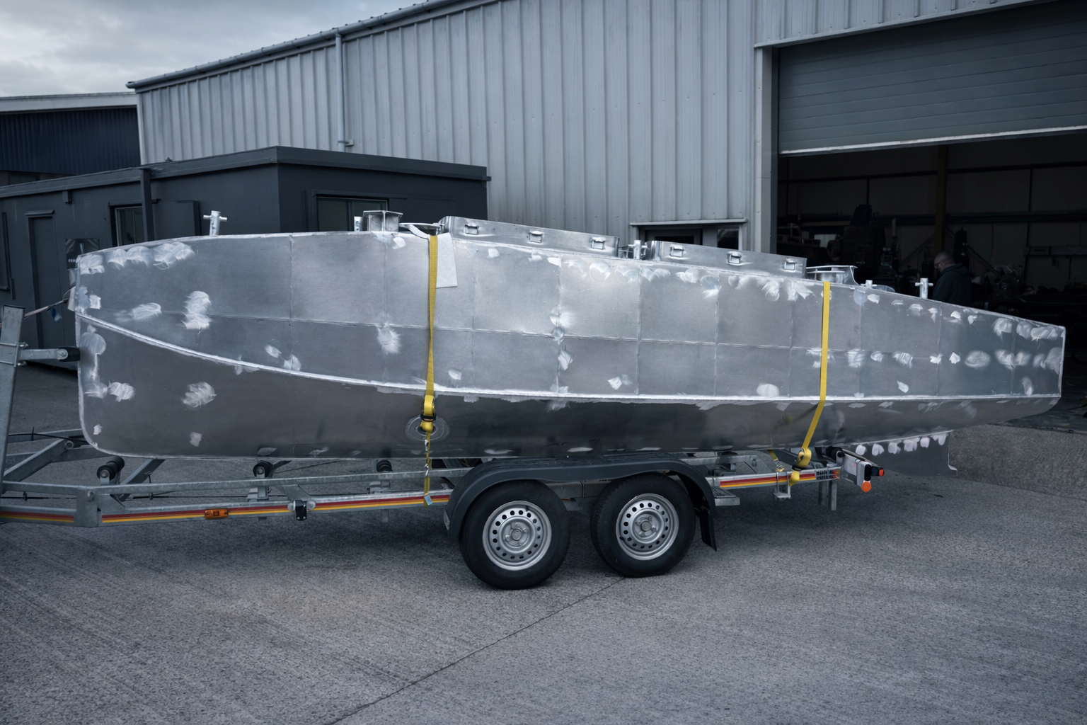 Unfinished aluminum boat on a trailer outside a warehouse, with visible weld marks and secured with yellow straps.