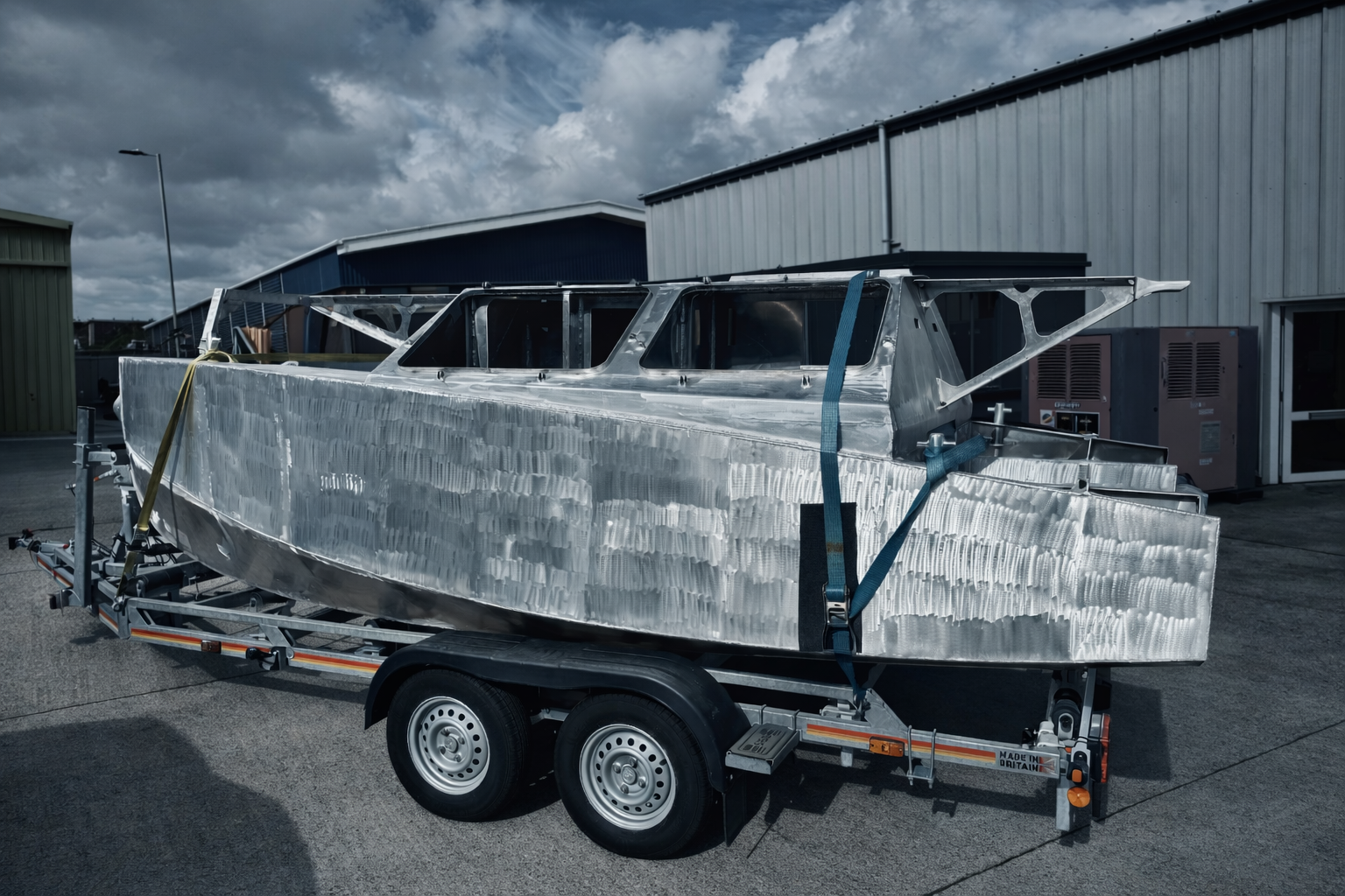 Unfinished boat made of aluminum on a trailer outside near industrial buildings, with dark clouds overhead.