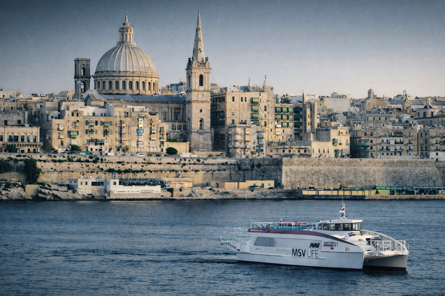 A passenger ferry on the water with a city skyline featuring historic buildings and domed church in the background.