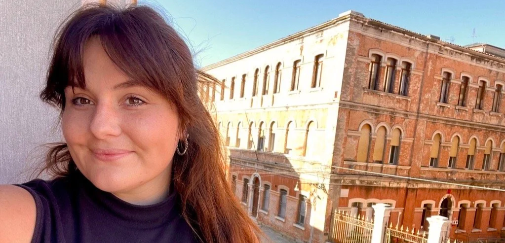 A woman with shoulder-length brown hair and hoop earrings is smiling for a selfie against a cityscape with a brick building in the background.