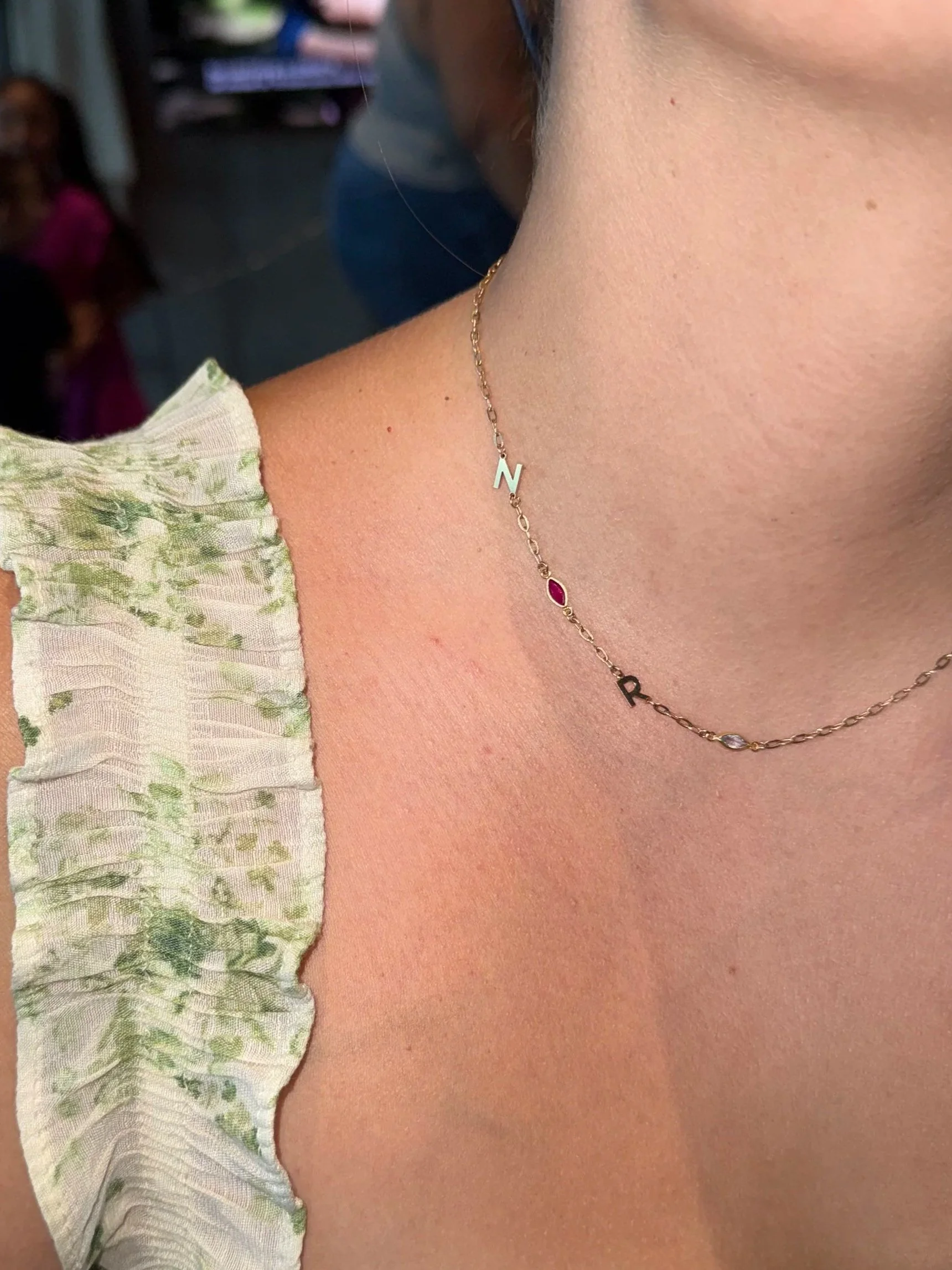 Close-up of a woman’s neck wearing a permanent 14k gold chain necklace with letter and gemstone charms, and a light-colored, floral-patterned ruffled sleeve.