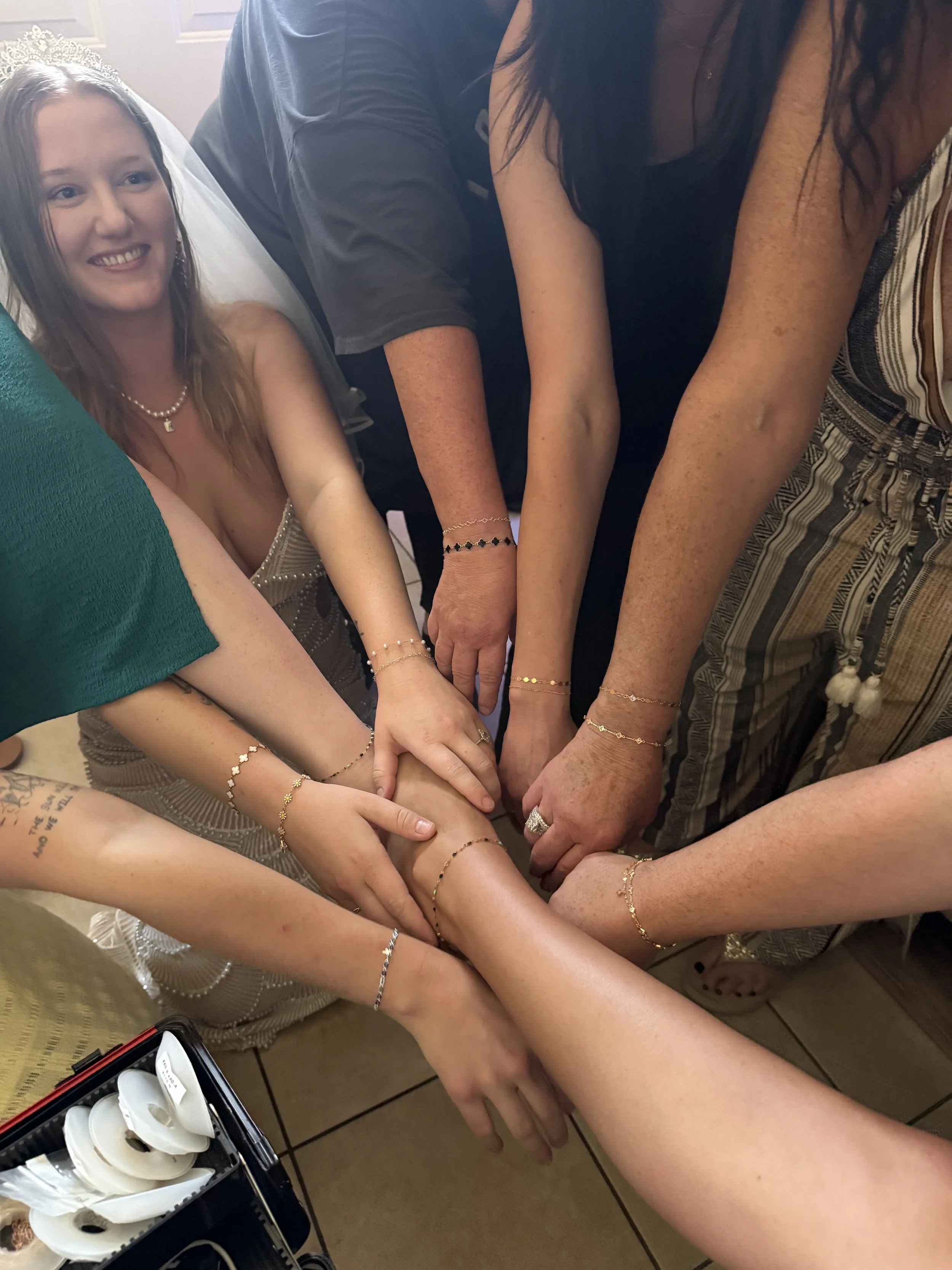 Multiple hands with various skin tones stacked together, each wearing a permanent bracelet, with a smiling girl in a wedding dress and tiara in the background on the left.