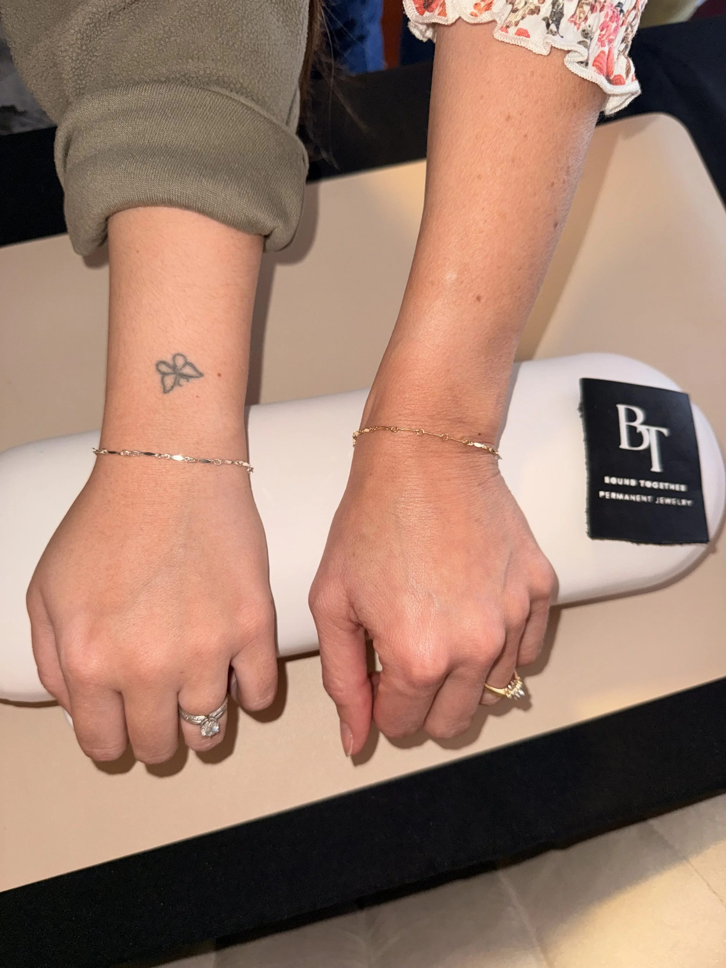 Two hands resting on a permanent jewelry display table. The left hand has an engagement ring with a large center stone and a gold bracelet. The right hand has a gold bracelet and a gold ring. The background includes permanent jewelry tags