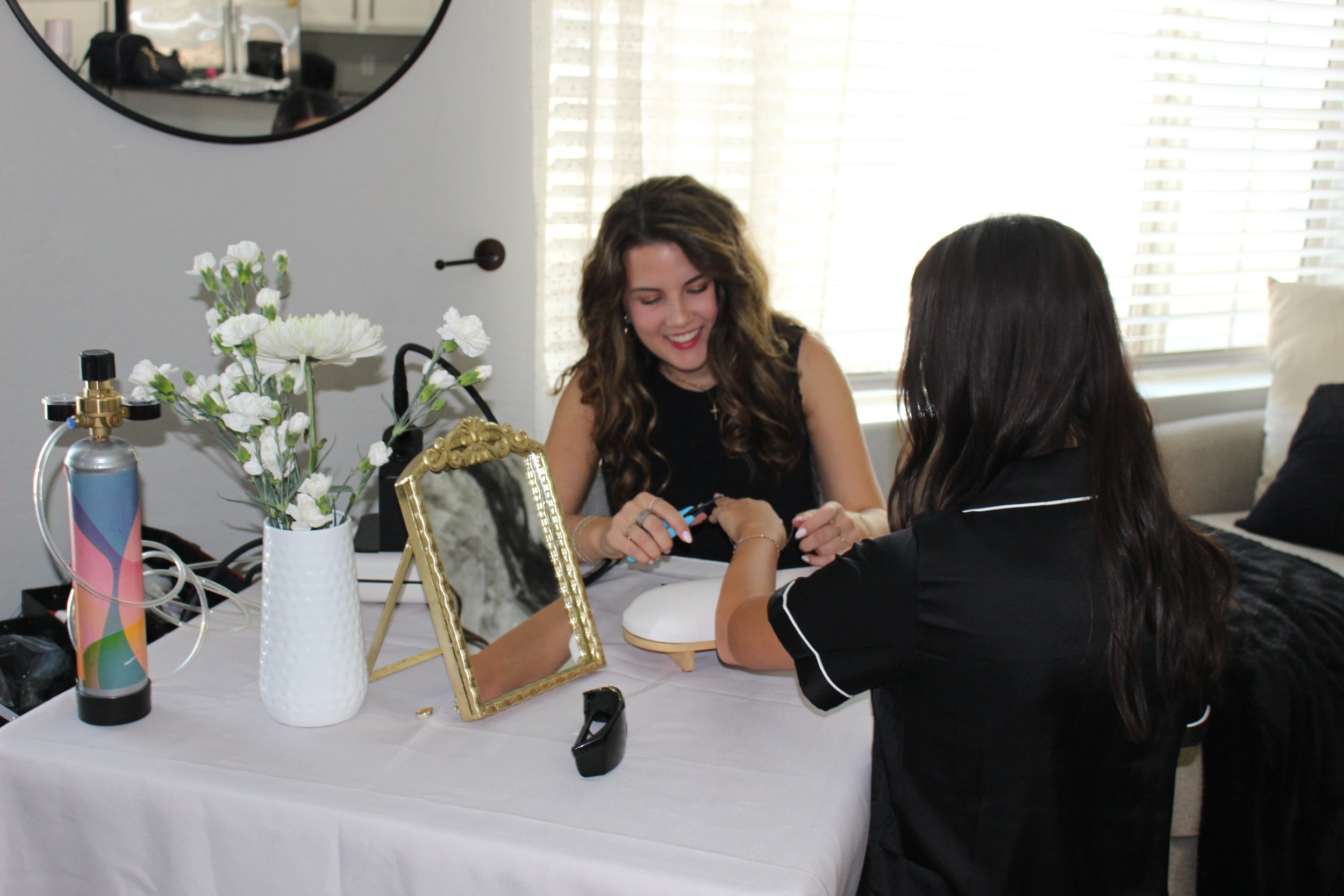 A woman with long curly brown hair and a black sleeveless top smiling as she receives a manicure from a woman with long straight dark hair wearing a black shirt with white trim, seated at a table with various decorative items including flowers in white pots, a gold-framed mirror, and nail tools, with a bright window with blinds in the background.