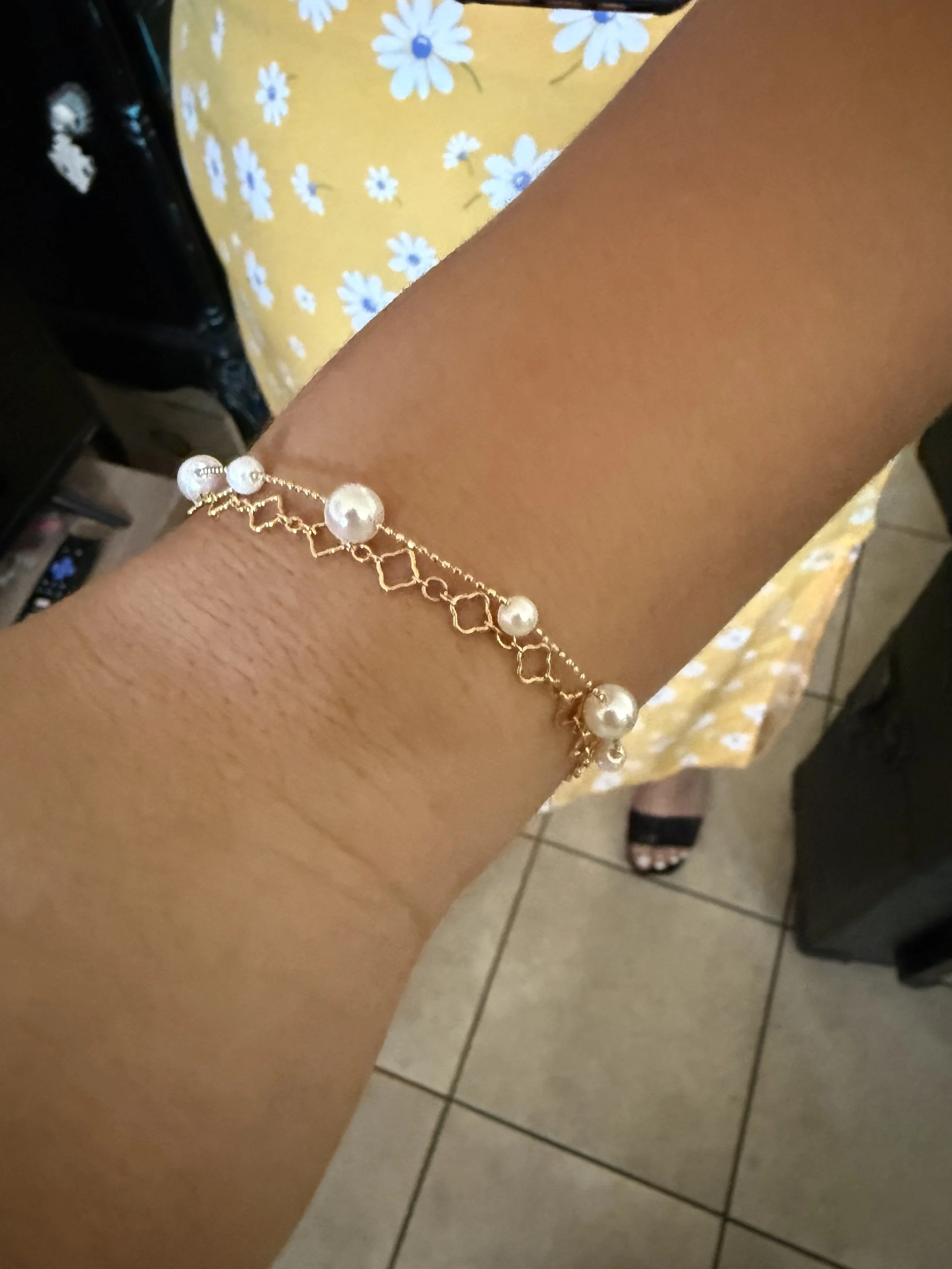 A person's wrist wearing a permanent gold bracelet with pearl and diamond accents, sitting next to a yellow fabric with white daisy flower pattern on a tiled floor.