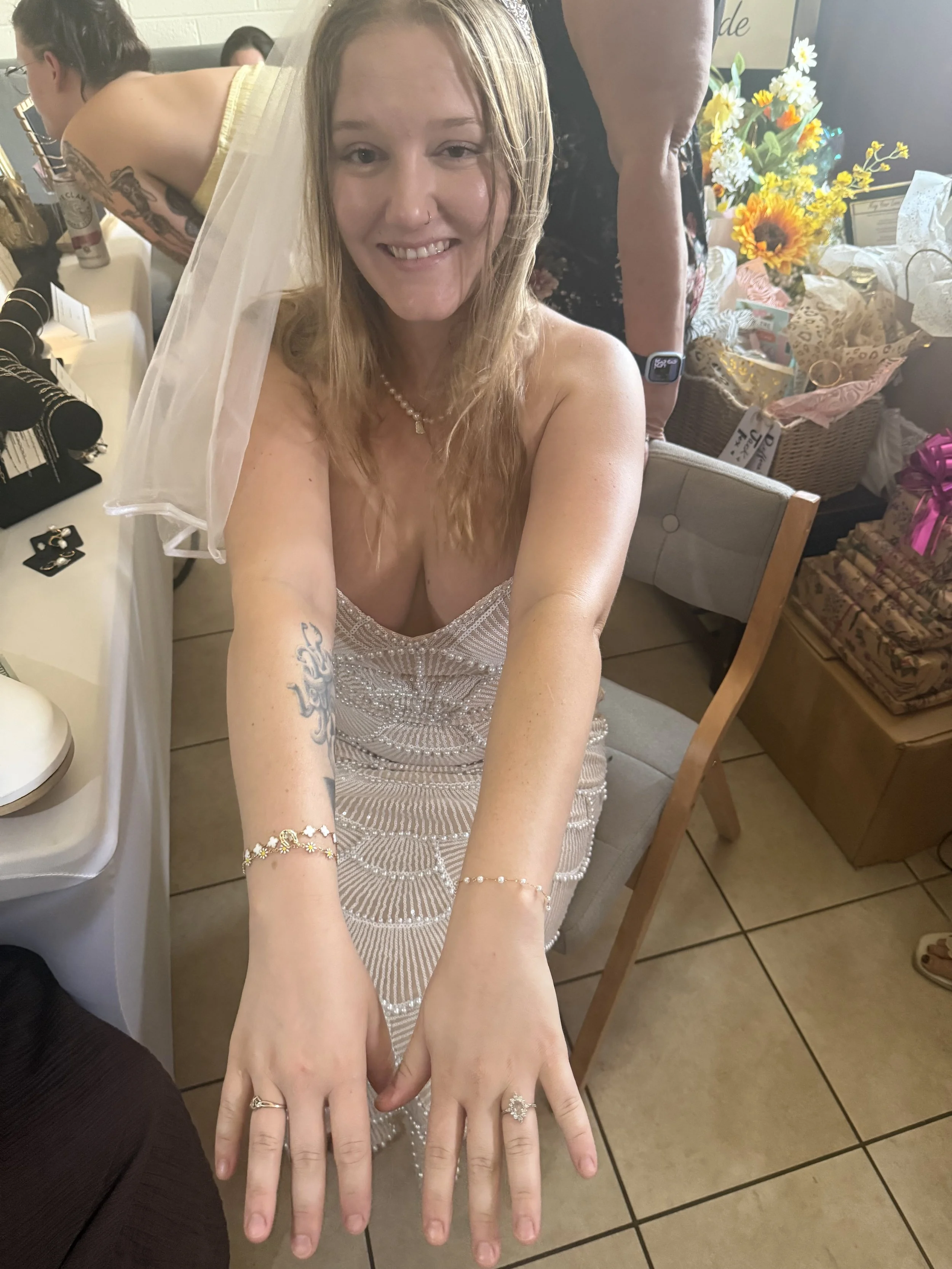 A woman with light brown, curly hair wearing a veil and a sleeveless dress, smiling at the camera. She has tattoos on her left arm and for rings on her fingers. In the background, there are flowers in a basket, a table with various items, and other p