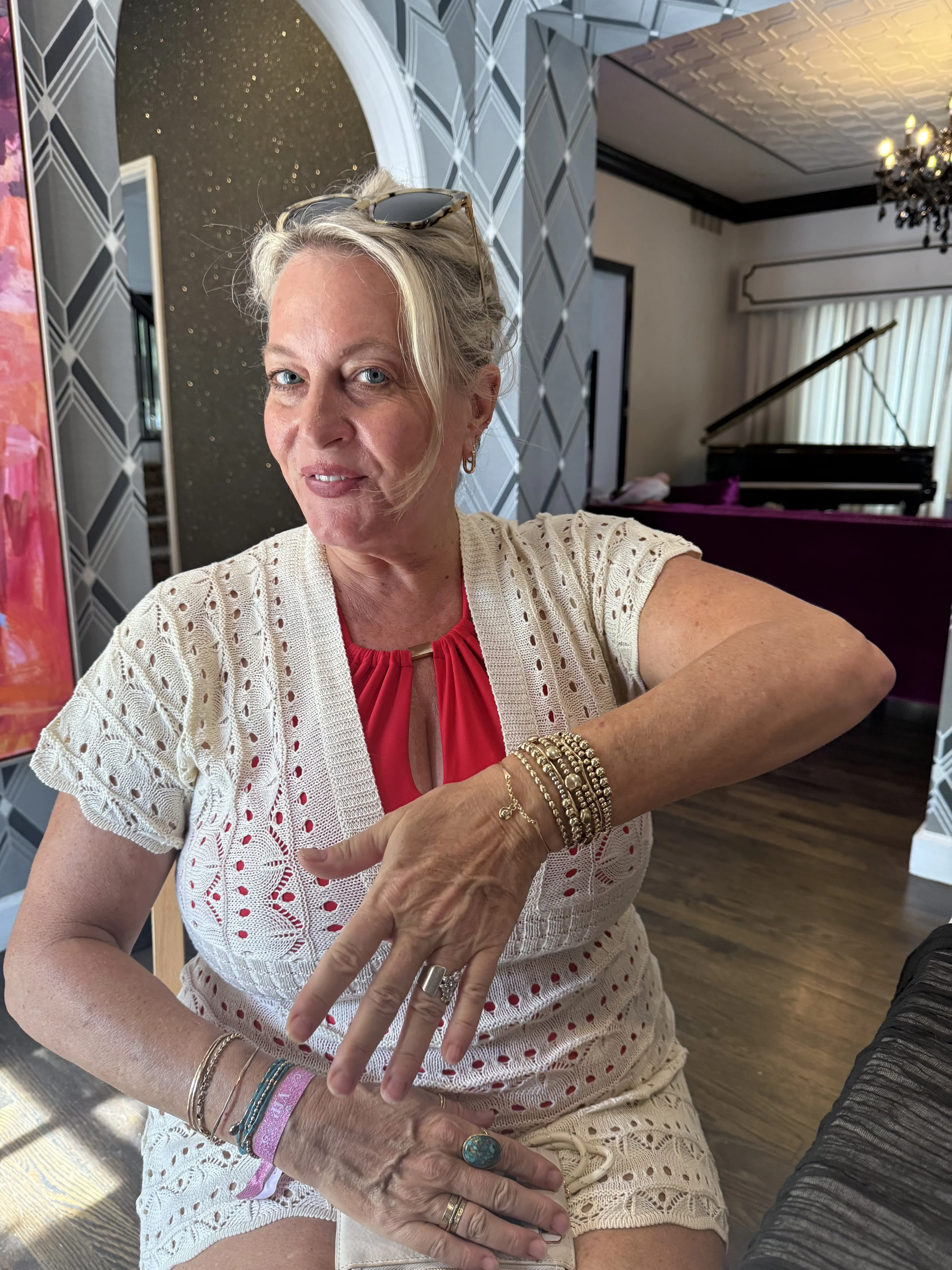 A woman with blonde hair, wearing sunglasses on her head, gold earrings, multiple bracelets, and a large turquoise ring, is taking a selfie. She is wearing a white, crochet-style top over a red shirt. She is sitting indoors near a dark purple grand piano, with a chandelier, a window with curtains, and patterned wallpaper visible in the background.