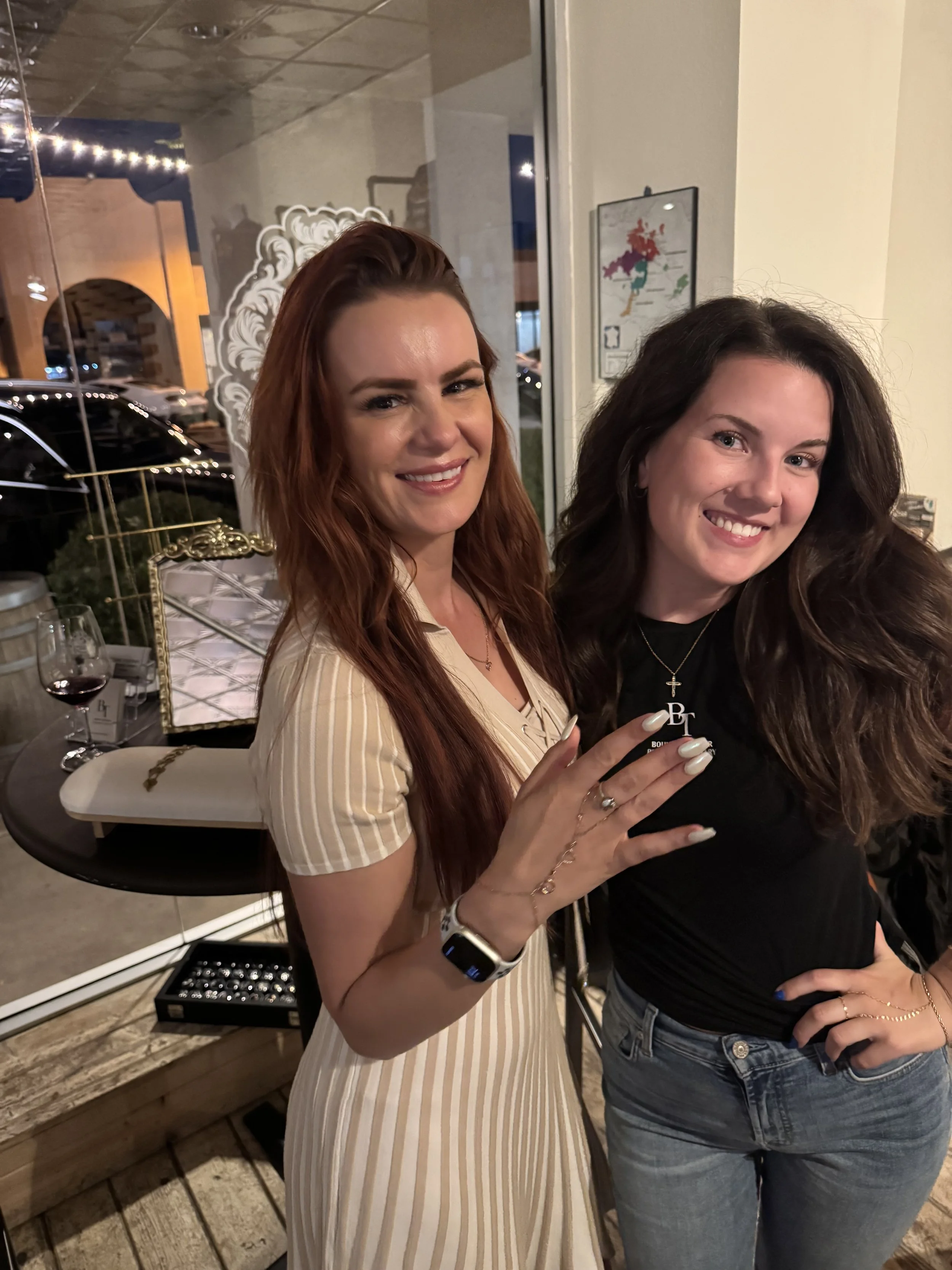 Two women smiling and standing close together in a restaurant. One has long, wavy red hair and is wearing a cream-colored, striped dress. The other has long, wavy dark hair and is dressed in a black top and jeans. A table with wine glasses and a jewelry box are visible behind them.