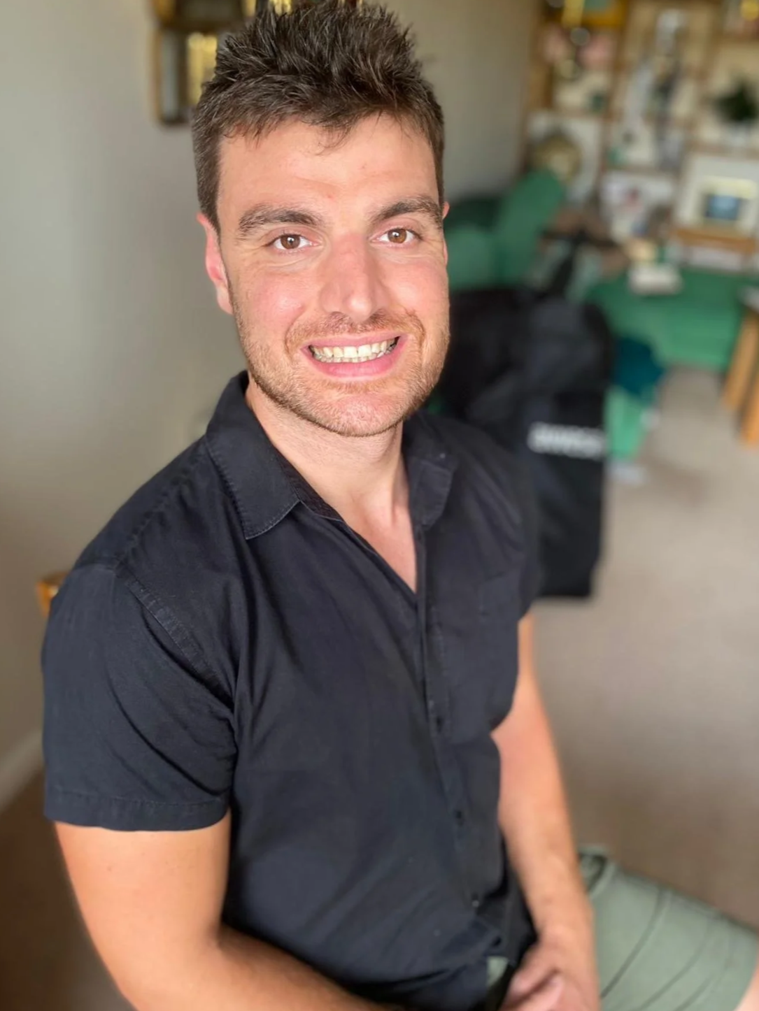 A smiling man with short brown hair wearing a black button-up shirt in a room with bookshelves and a green chair in the background.