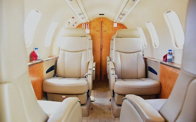 Interior of a private jet with beige leather seats and small side tables, water bottles, and windows on each side.