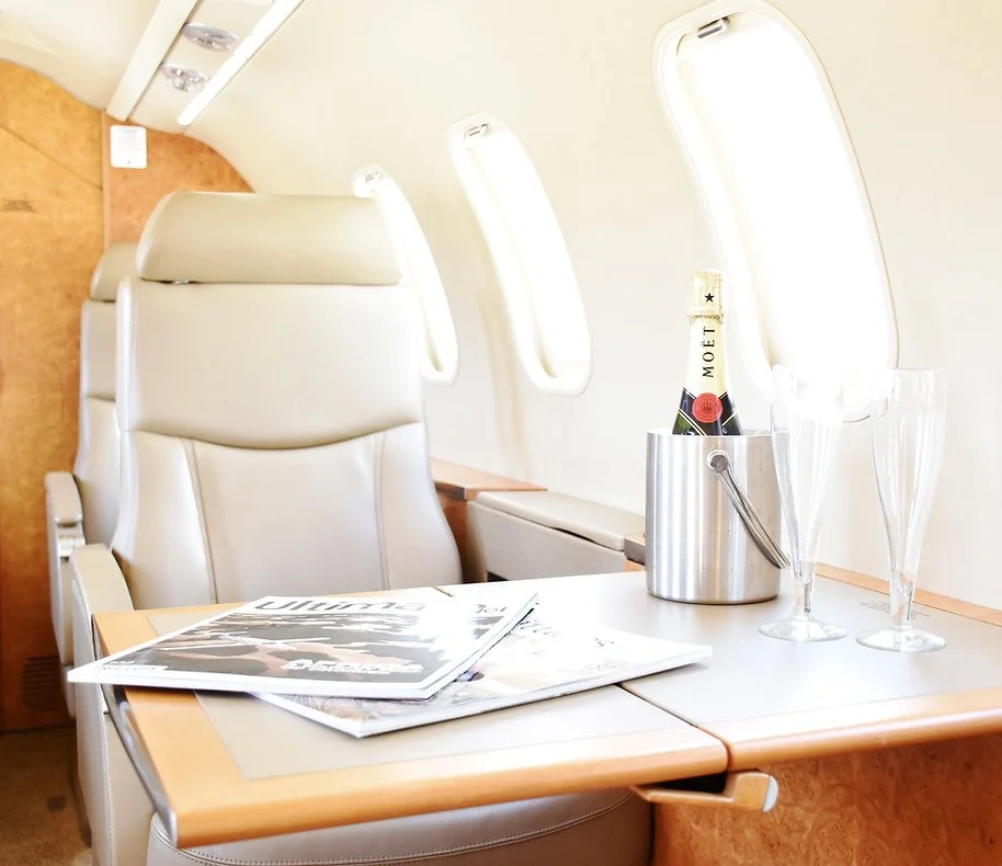 Luxury airplane seat with a wooden tray table holding magazines, a champagne bottle in an ice bucket, and empty champagne flutes.