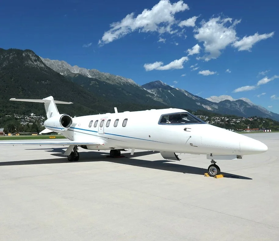White private jet parked on a runway with mountains in the background and a blue sky with scattered clouds above.