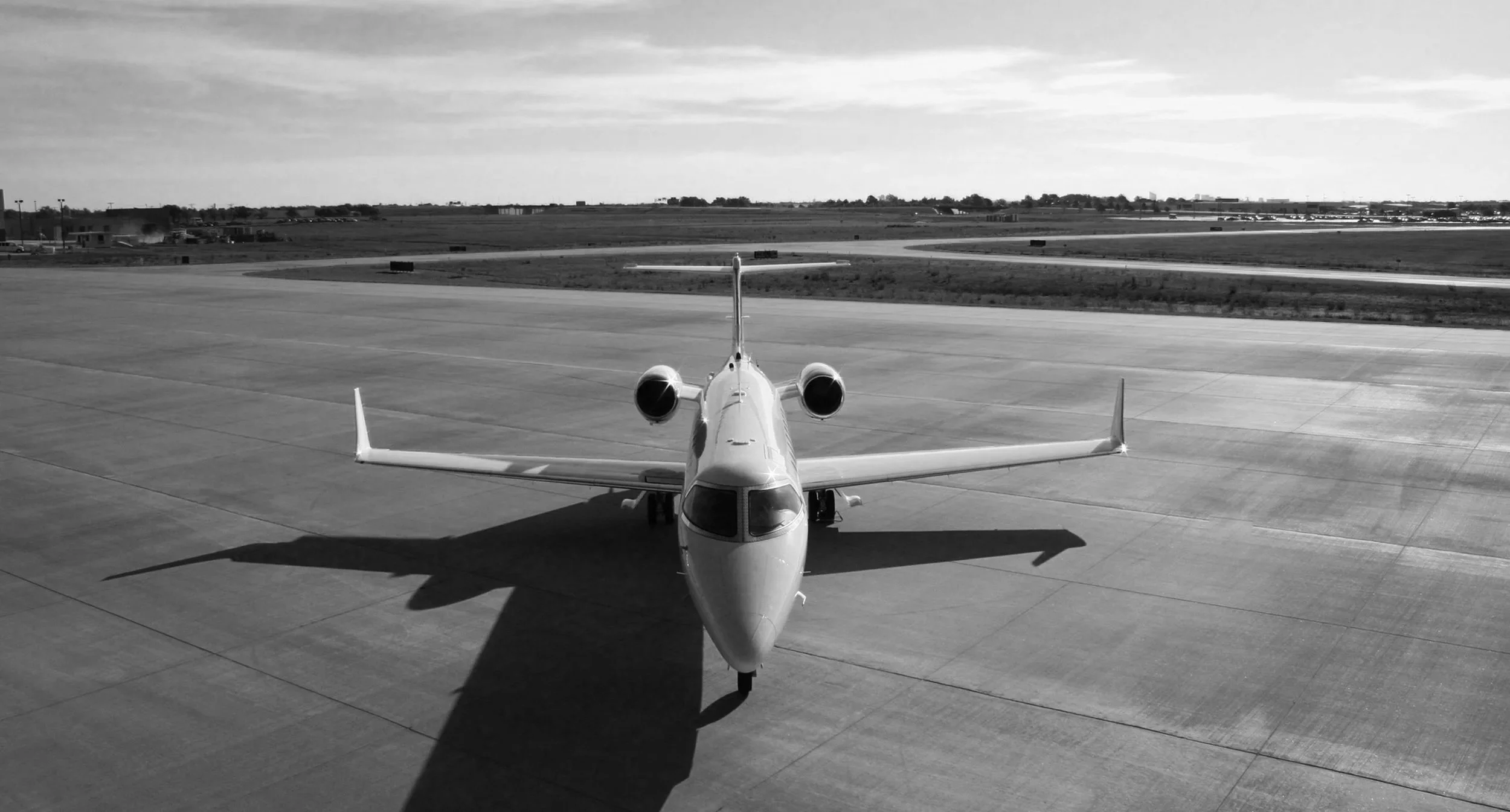 A private jet airplane on an airport tarmac, with long shadows cast on the ground, under a partly cloudy sky.