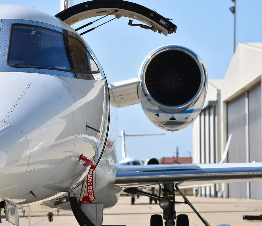 Close-up of a private jet airplane on the tarmac with the cockpit door open, showing the engine and landing gear, under a clear blue sky.