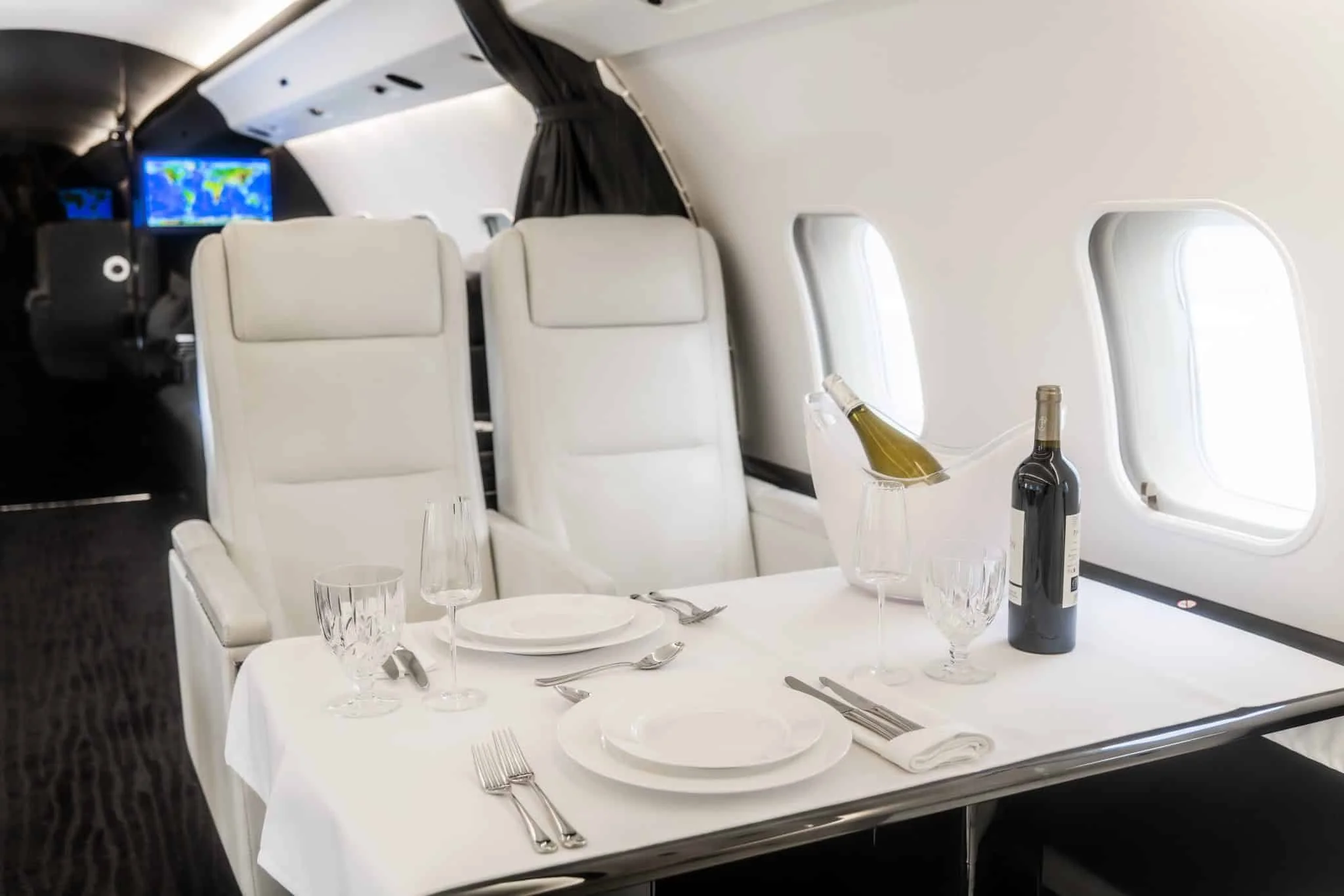 Luxury airplane dining area with a table set for two, including two white plates, silverware, wine glasses, a bottle of wine, and a bucket with a bottle of champagne, inside a private jet with white seats and windows.
