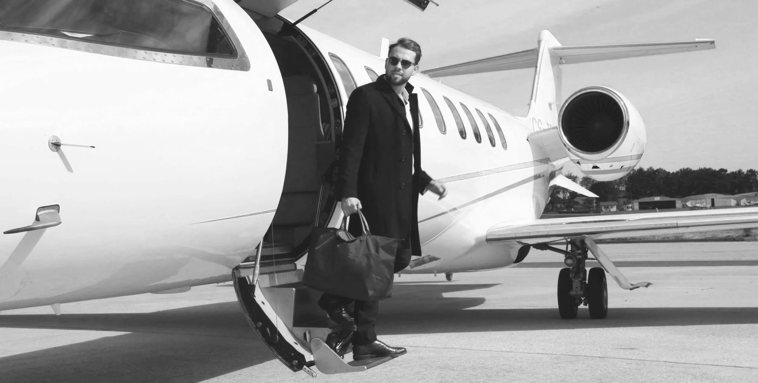 A man in sunglasses and a long coat carrying a handbag boarding a private jet on runway.