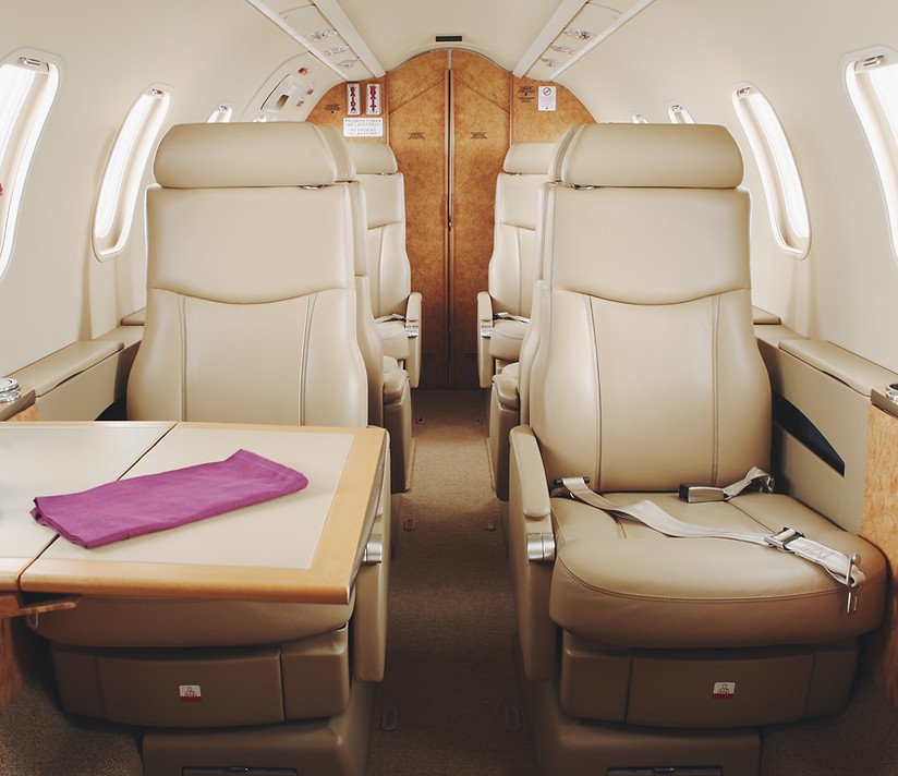 Interior of a private airplane cabin with beige leather seats, a wooden table with a purple cloth, and windows along the side wall.