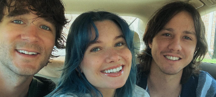 IMY2. Three friends smiling in a car, two men and a woman with blue hair, all looking at the camera. band.