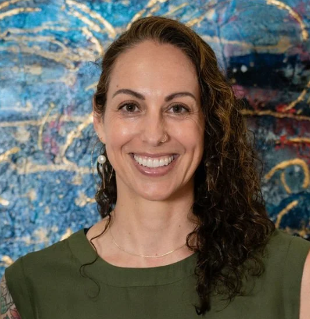 A woman with curly hair and a nose ring smiling in front of a colorful, abstract background. Courtney Trowman. cofounder of AURi LLC