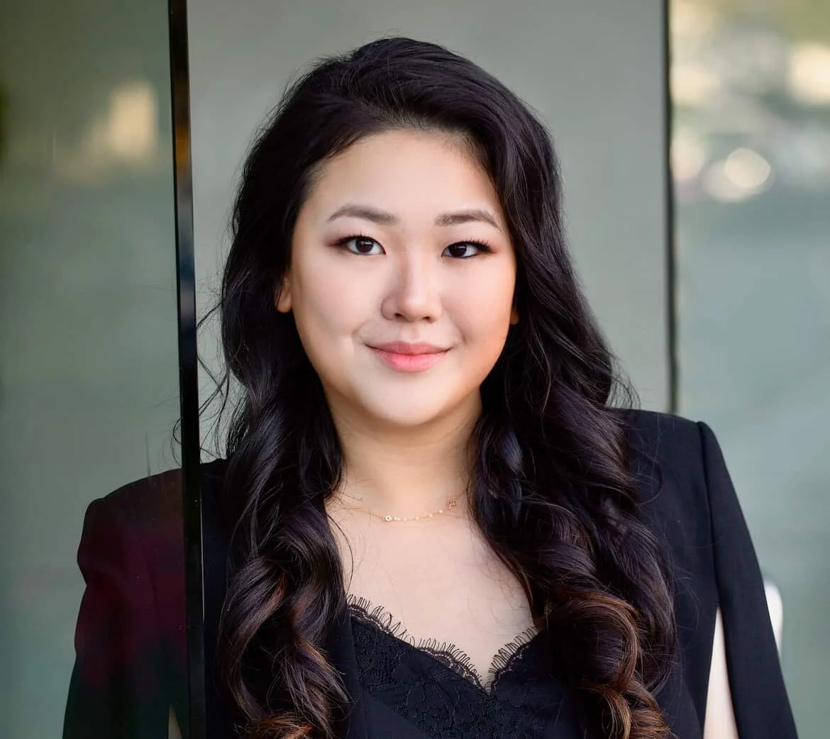 A young woman with long, wavy black hair, wearing a black blazer and a lace top, smiling outdoors with a blurred background. Nat Z.