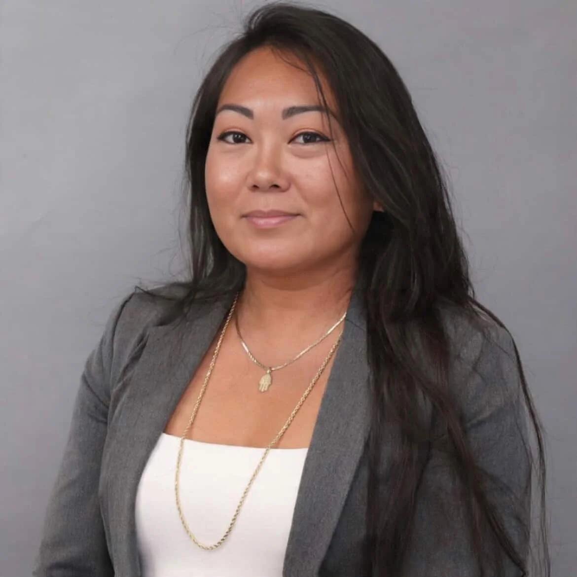 A woman with long black hair, wearing a gray blazer and white top, smiling subtly against a gray background, accessorized with layered necklaces. Cookie S.
