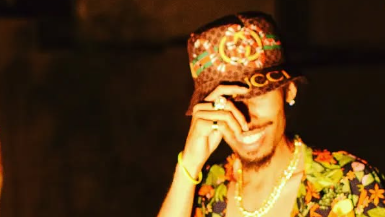 A man wearing a colorful cap, floral shirt, and gold chain, smiling with his hand near his face. Rapper Kush Kloudz