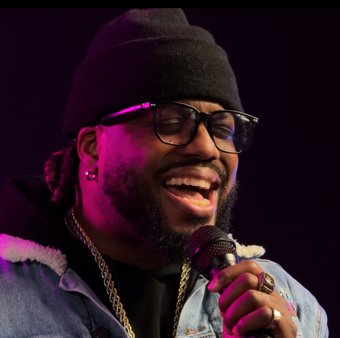 A man with glasses, a black beanie, and hoop earrings is singing or speaking into a microphone, smiling, with purple lighting on him.