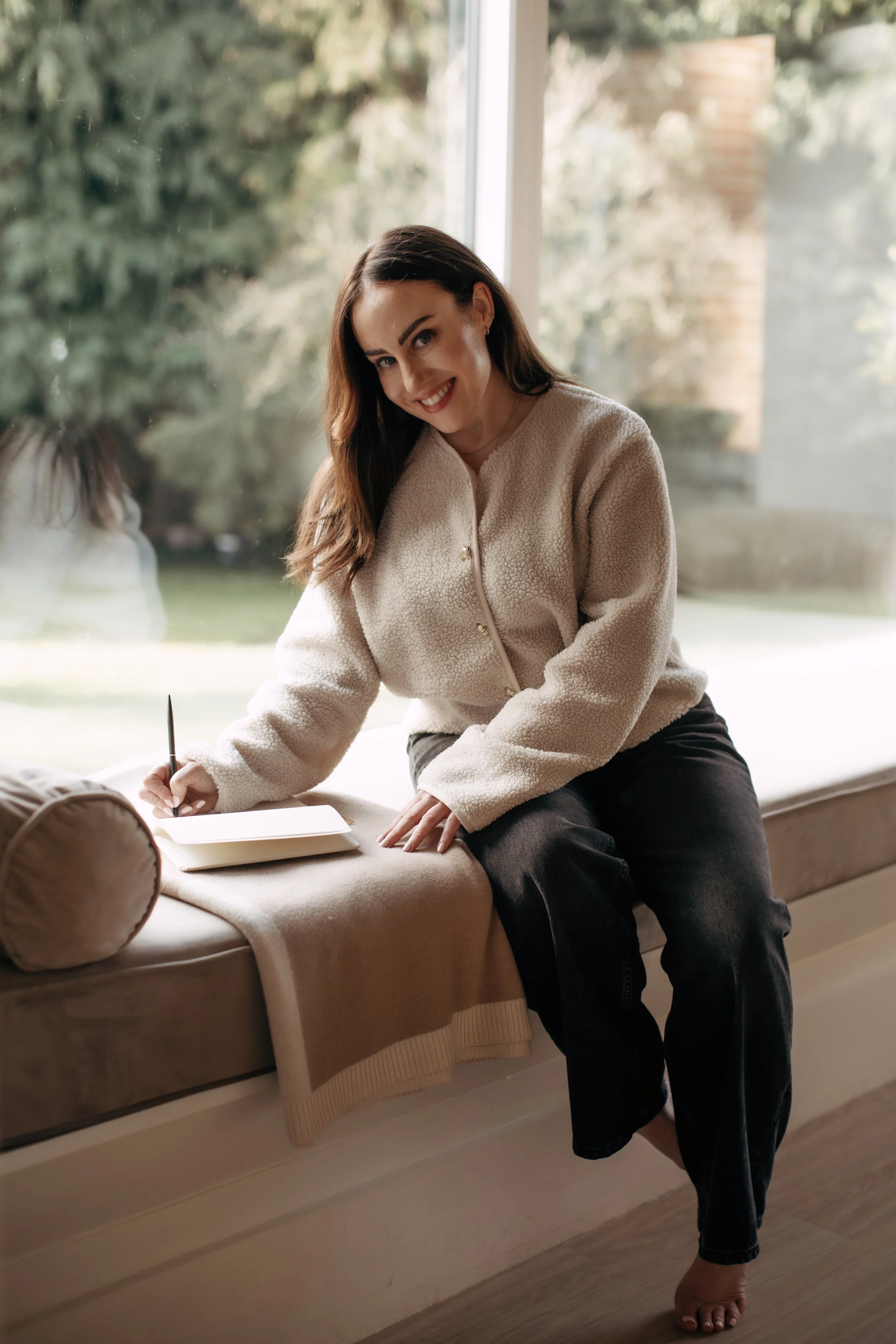 A woman with long brown hair, wearing a cream-colored sweater, sitting on a window seat, smiling, writing in a notebook with a pen, near a large window with greenery outside.