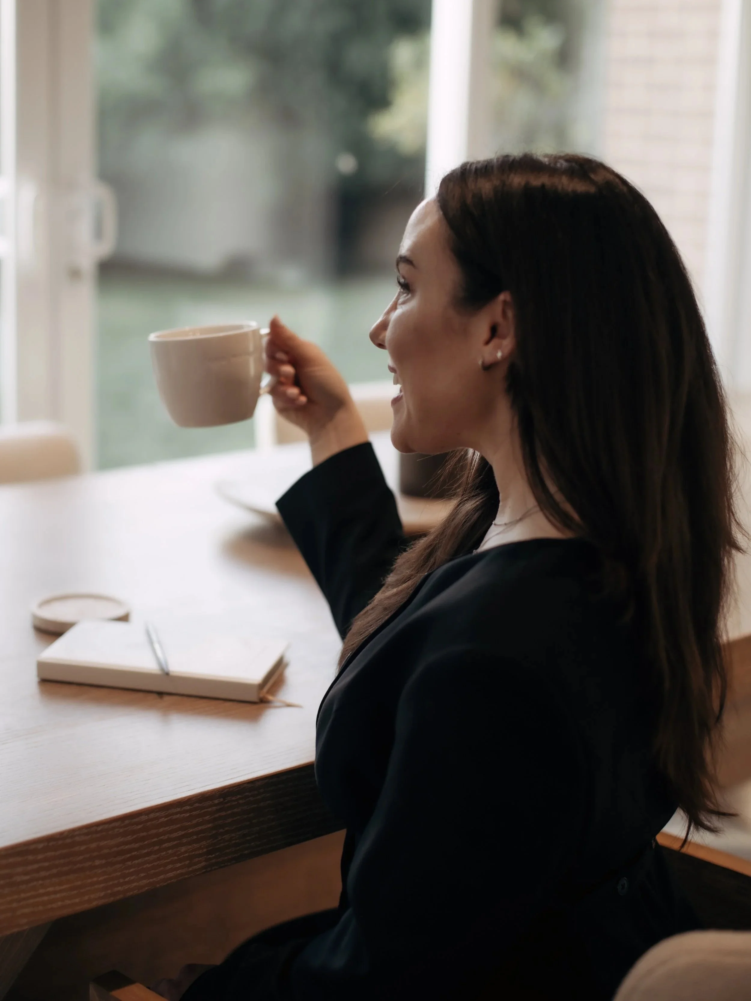 A woman with long dark hair, wearing a black jacket, sitting at a wooden table, smiling and drinking from a beige mug near a window.