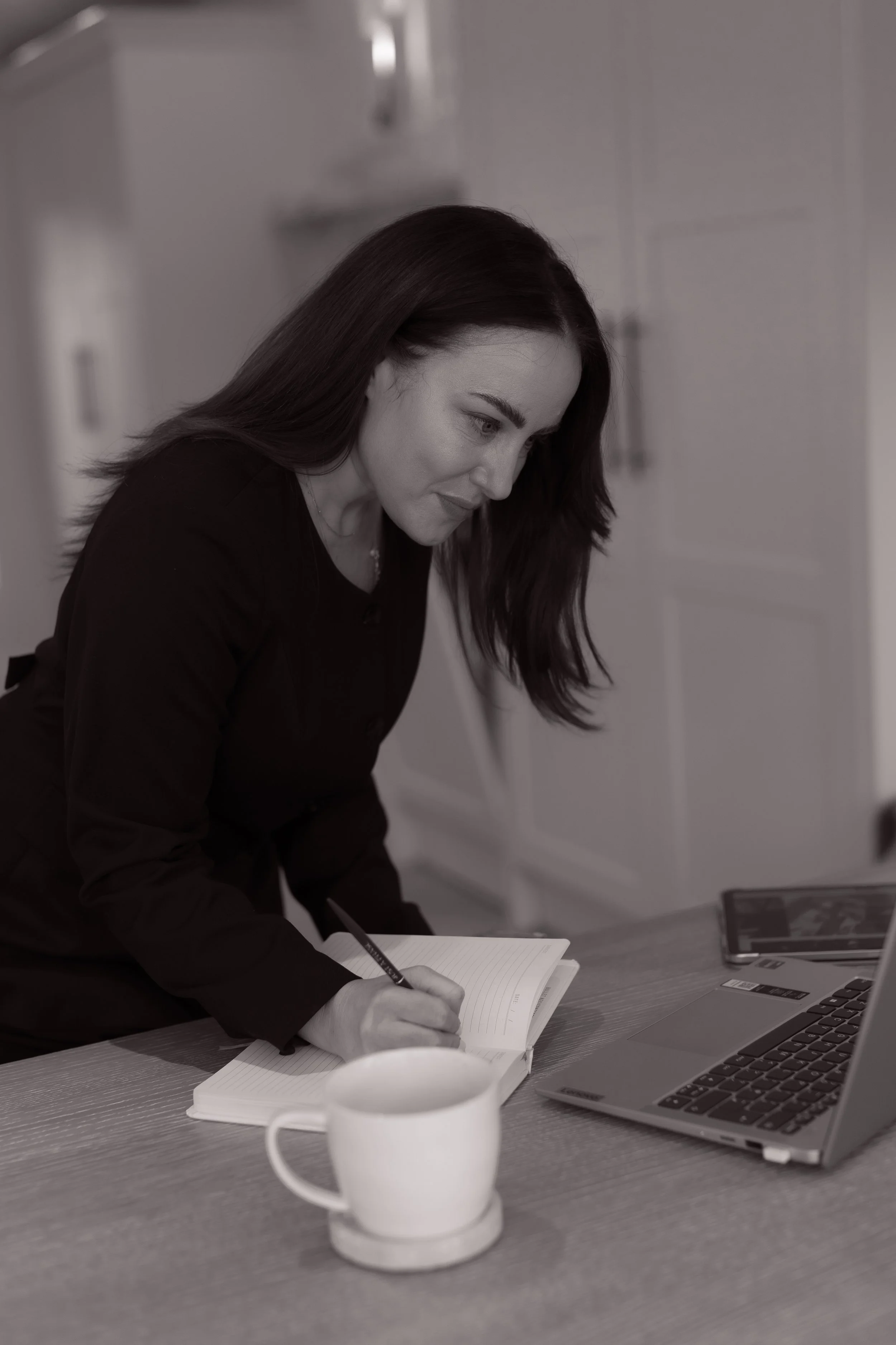 A woman with dark hair writing in a notebook at a table with a laptop, a mug, and a smartphone.