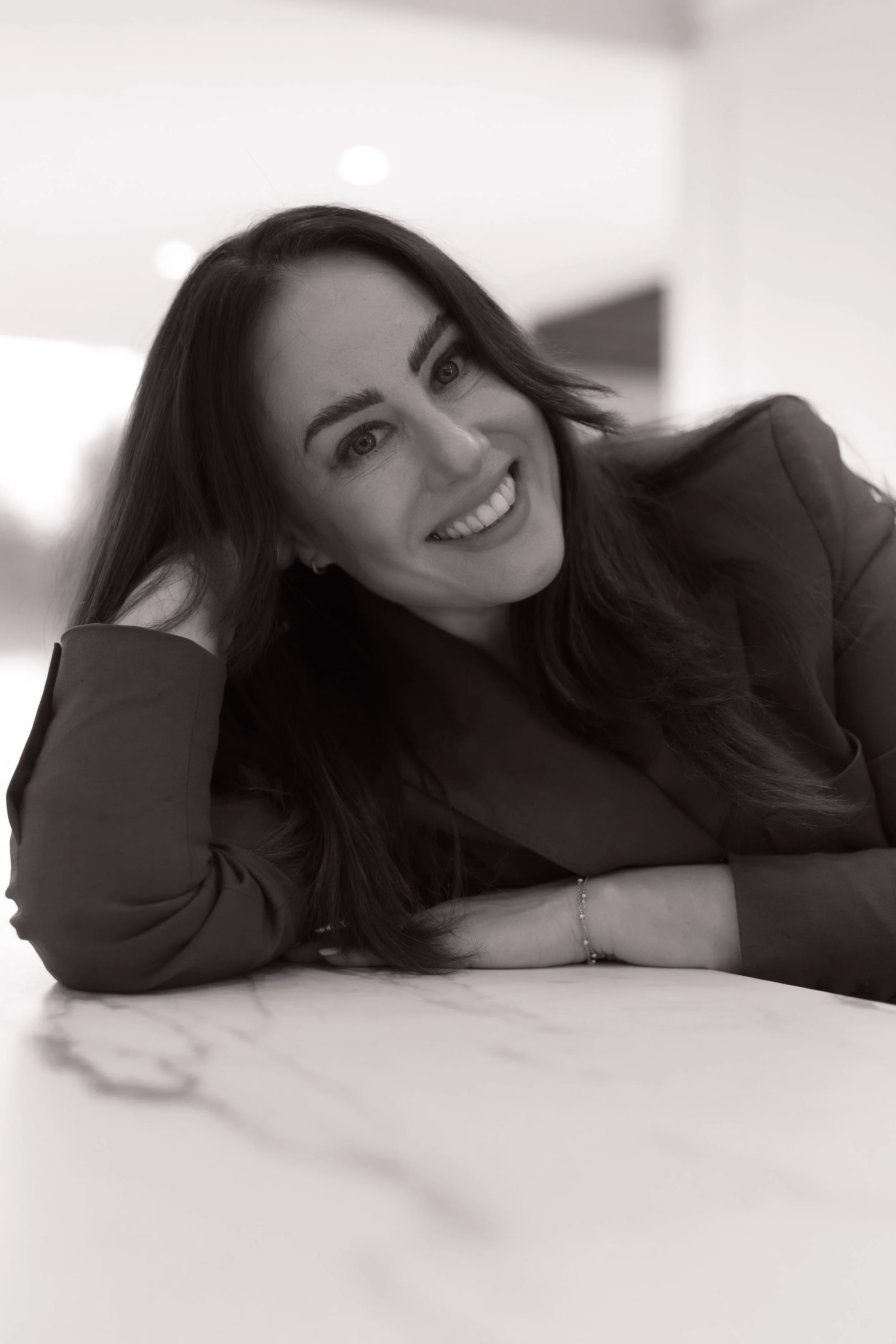 Black and white portrait of a woman relaxing with her head on her hand, smiling at the camera, with long dark hair, wearing a blazer, and a bracelet on her wrist.