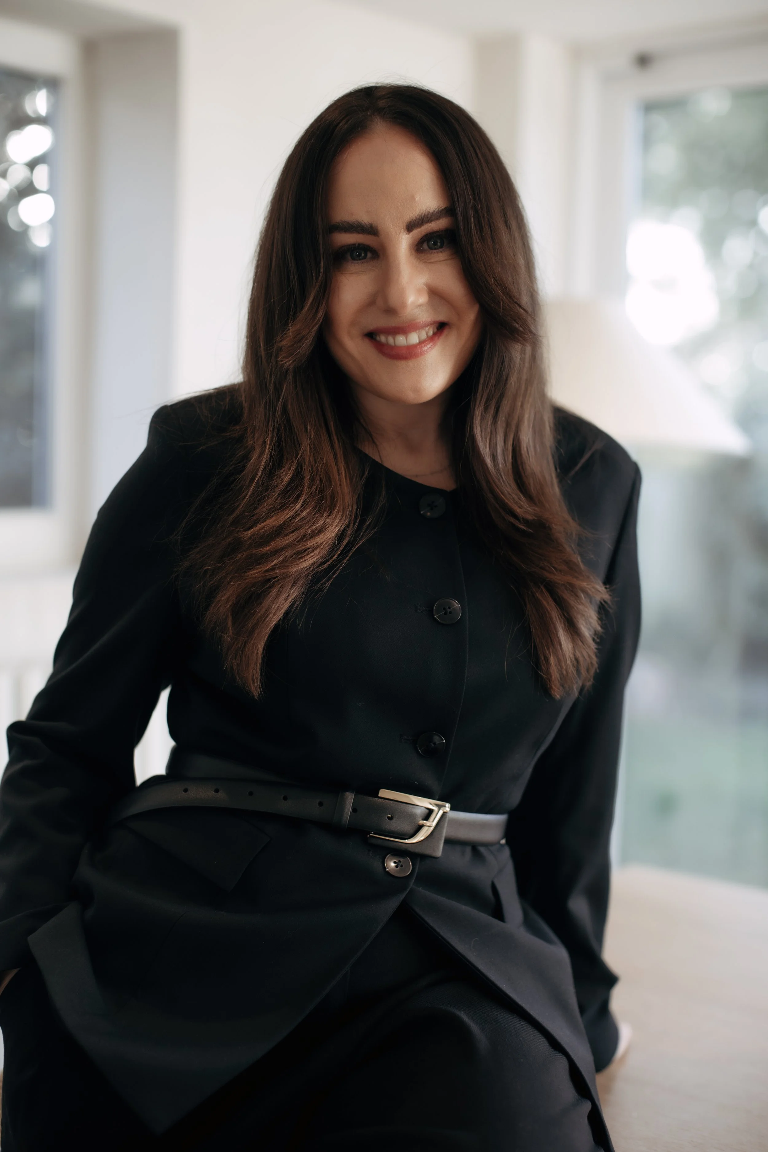A woman with long brown hair, wearing a black blazer and matching pants, sitting indoors with a blurred background of windows and greenery.