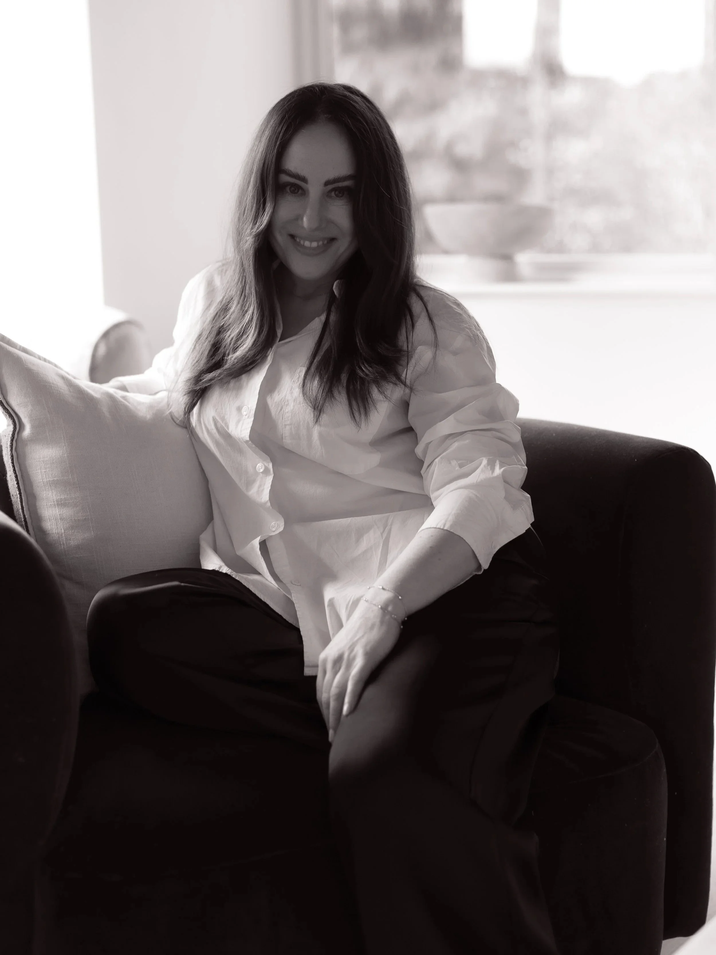 A woman with long, dark hair smiling while sitting on a dark sofa, wearing a white shirt and dark pants, in a well-lit room with a window in the background.