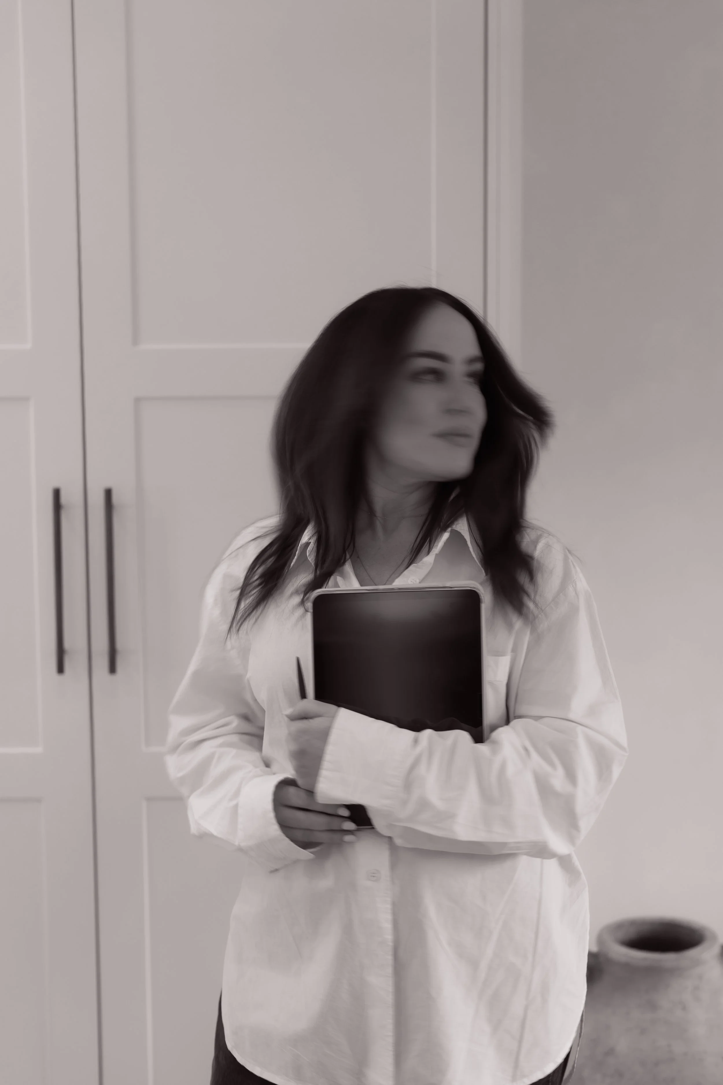 A woman with dark, shoulder-length hair holds a tablet or notebook, standing in a room with white cabinets and a tiled wall.
