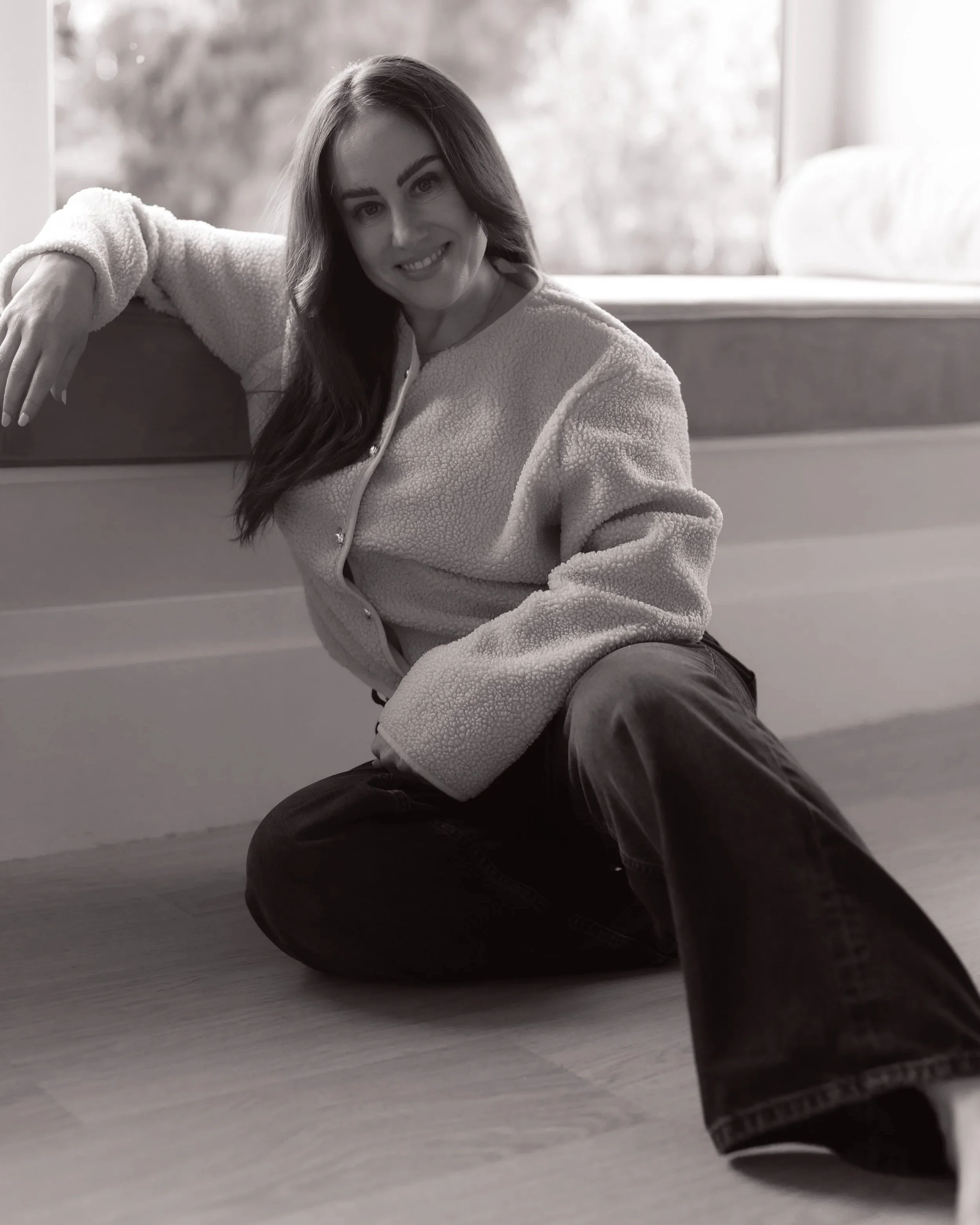 A woman with long hair sitting on the floor near a large window, smiling at the camera, wearing a light-colored fleece jacket and dark pants.